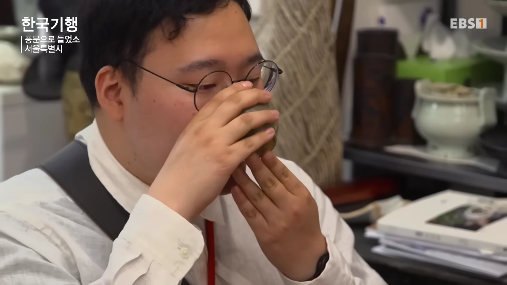 안경을 쓴 남성이 두 손으로 작은 찻잔을 감싸 쥐고 차를 마시고 있는 모습