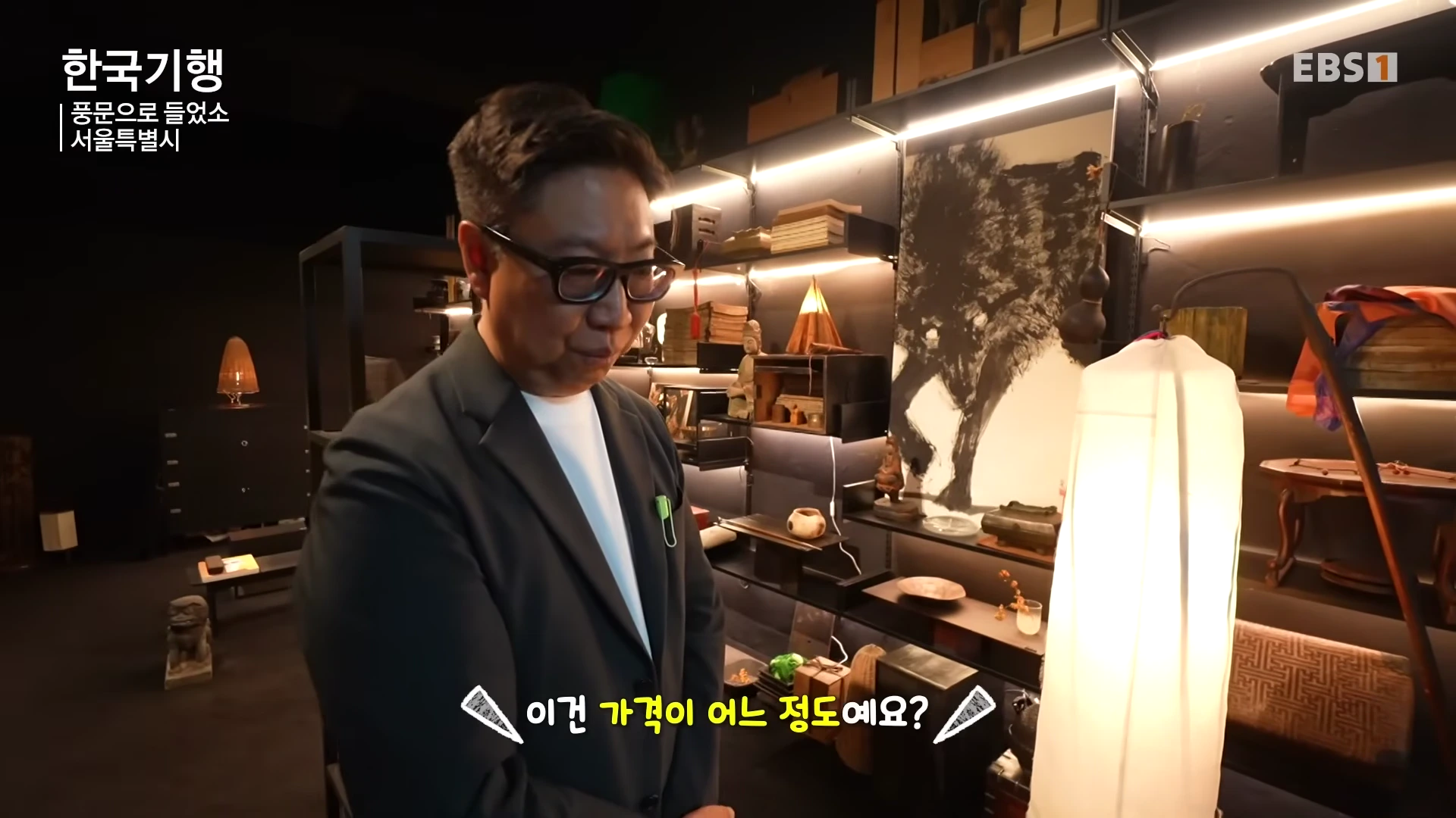 안경을 쓴 남성이 조명이 켜진 진열장 앞에 서서 골동품을 살펴보고 있는 모습