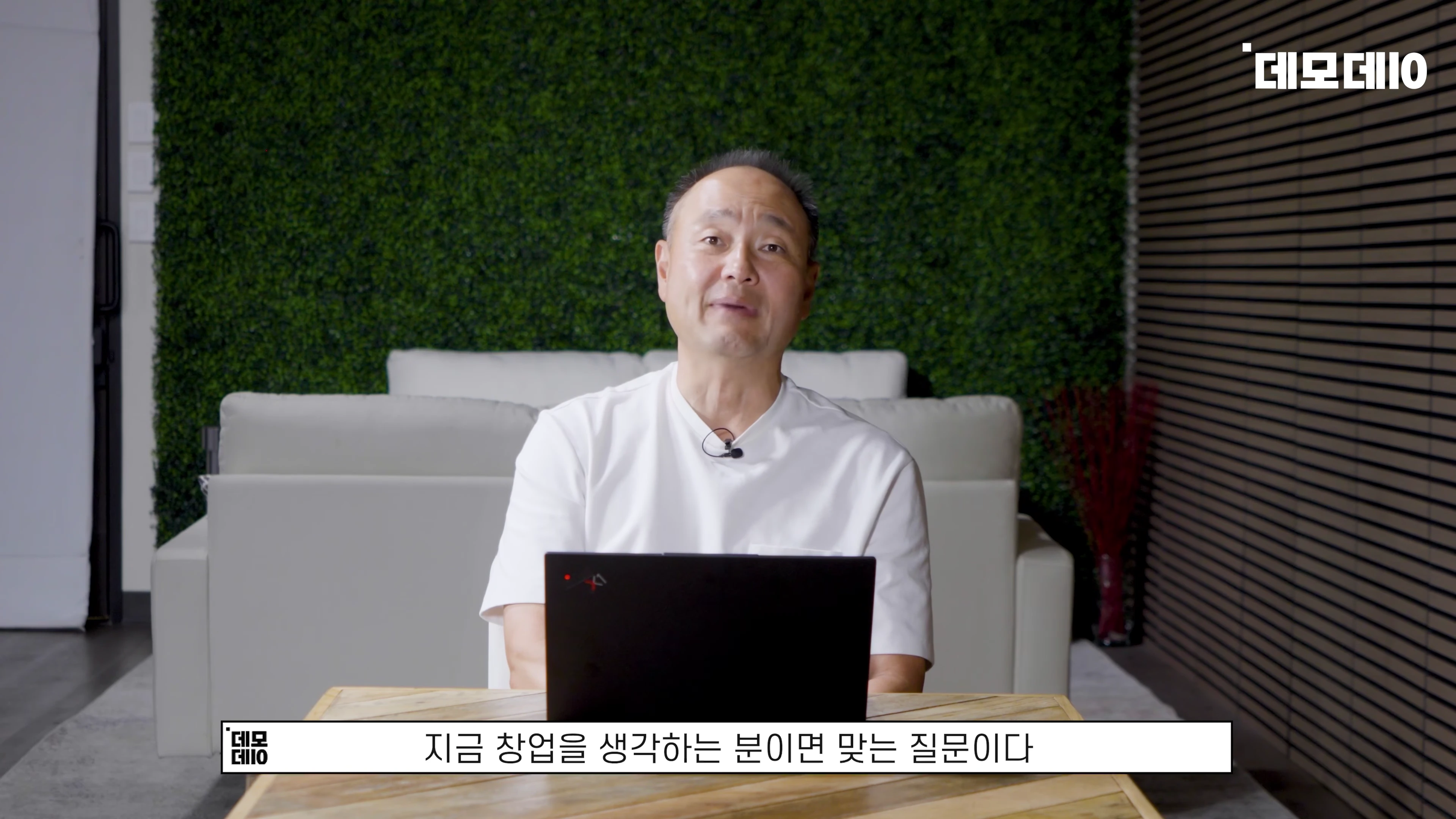 흰색 티셔츠를 입은 중년 남성이 책상 앞에 앉아 노트북을 보며 이야기하고 있다.