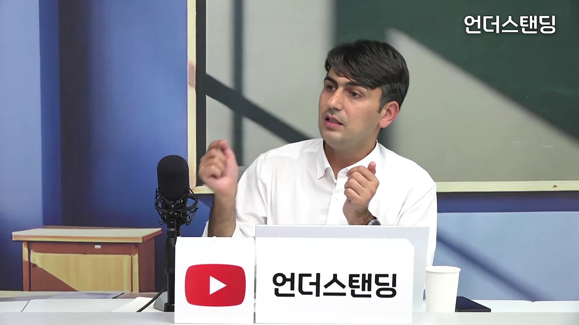 흰 셔츠를 입은 남성이 스튜디오에서 마이크 앞에 앉아 손짓하며 대화하고 있다.