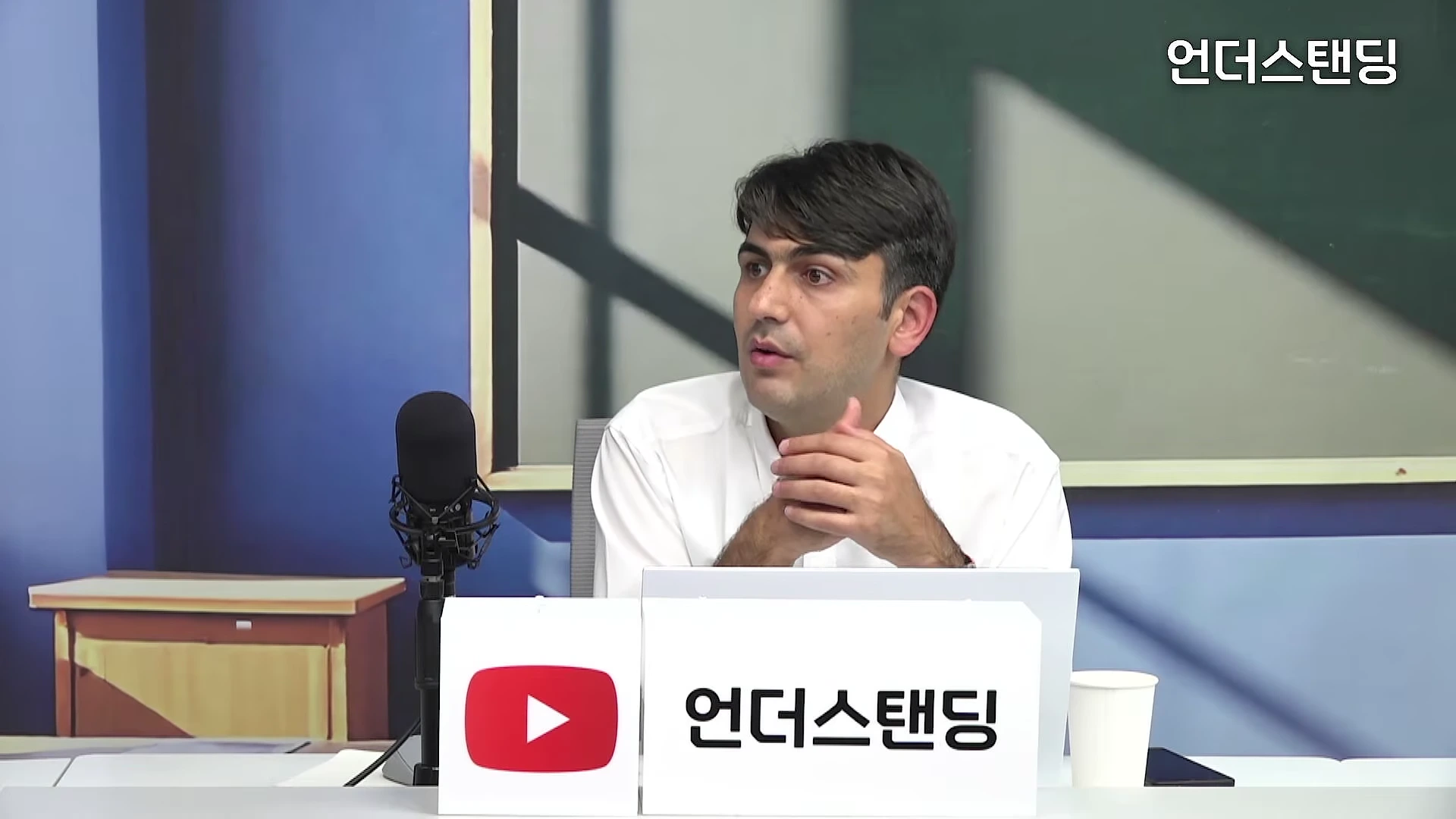 흰 셔츠를 입은 남성이 스튜디오 책상에 앉아 마이크를 앞에 두고 이야기를 나누고 있다.