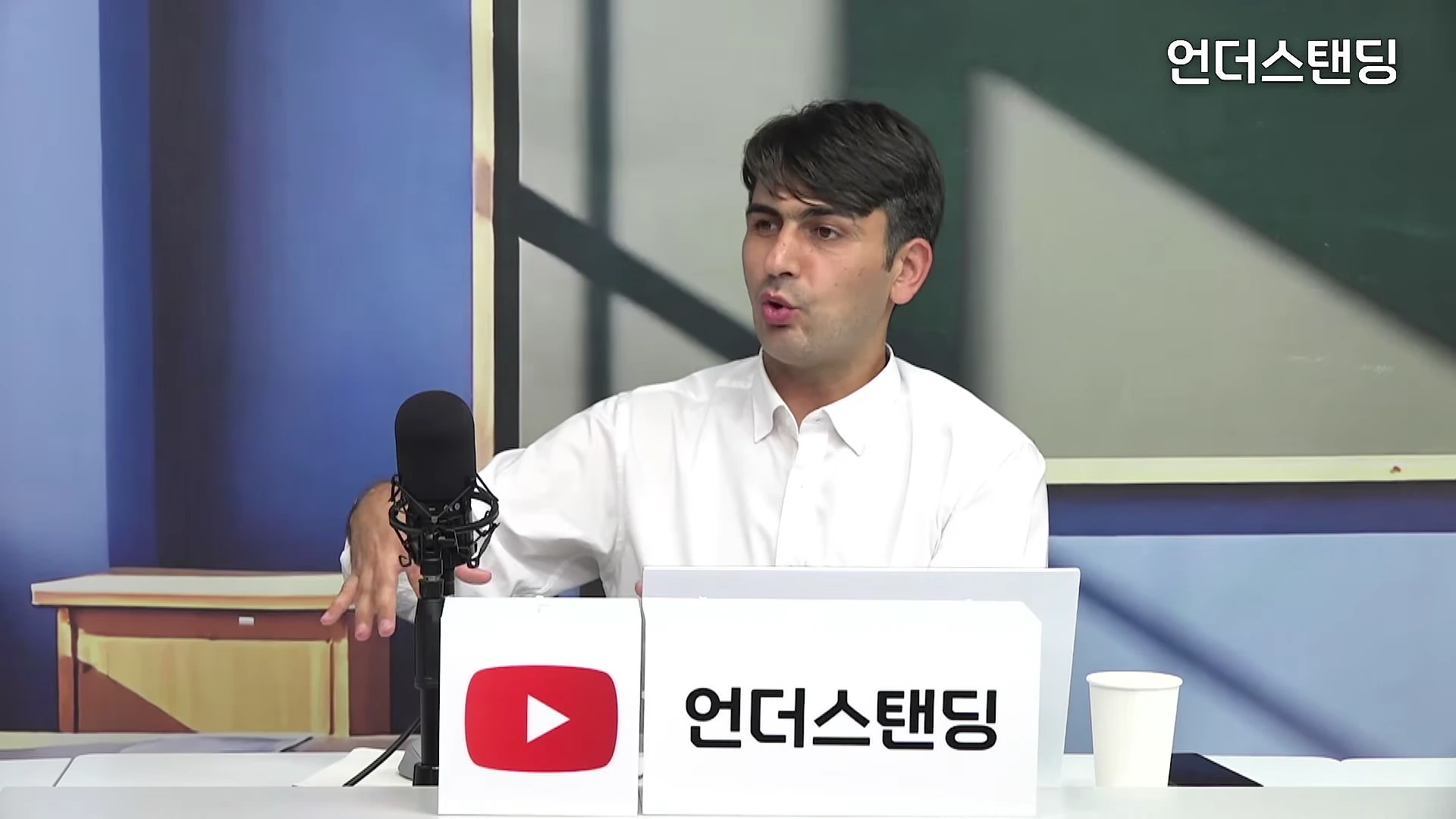 스튜디오에서 흰 셔츠를 입은 남성이 마이크 앞에 앉아 손짓하며 대화하고 있다.