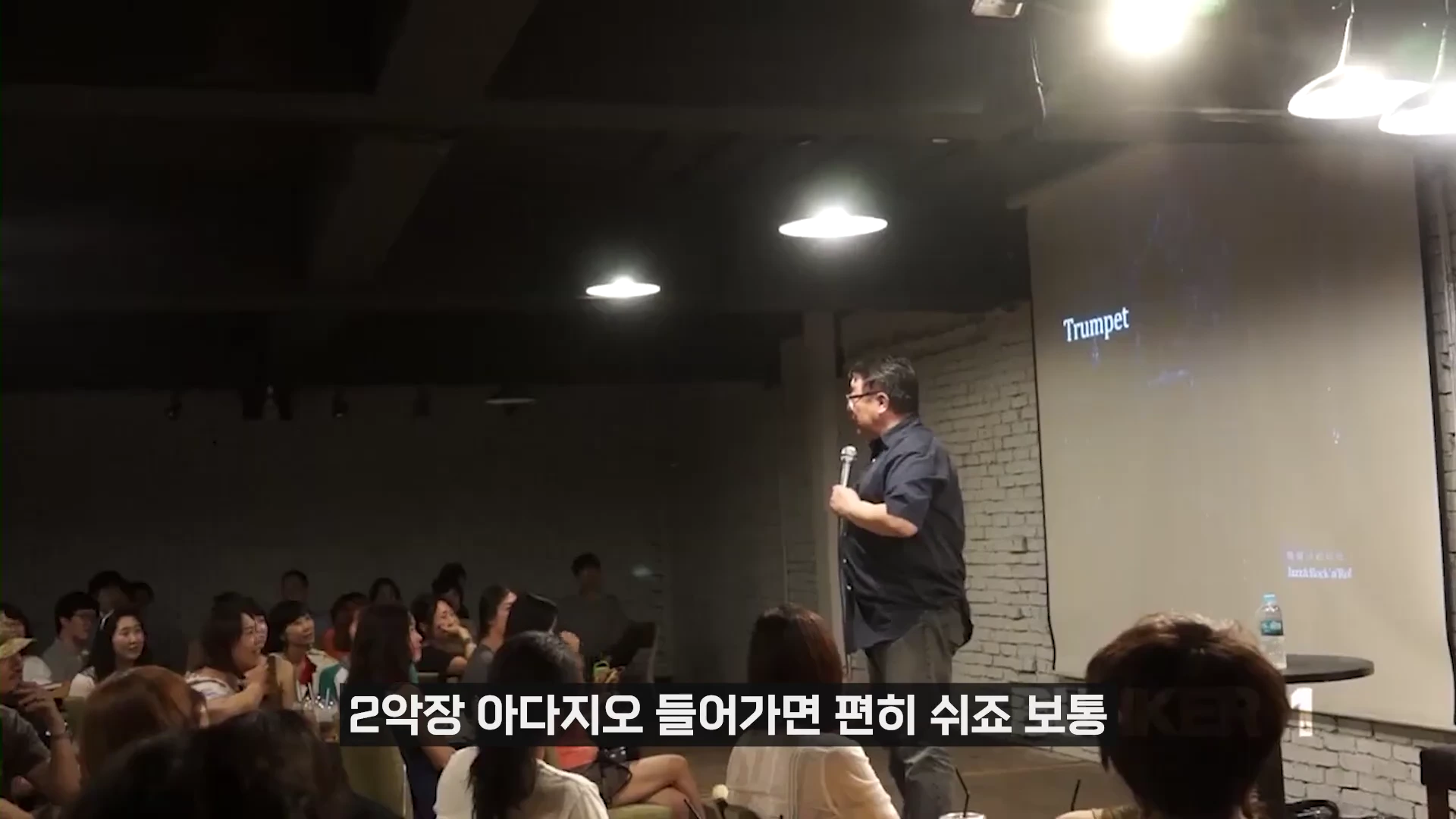 강연자가 어두운 실내에서 마이크를 들고 청중을 향해 이야기하고 있으며, 옆 스크린에는 'Trumpet'이라는 글자가 떠 있다.