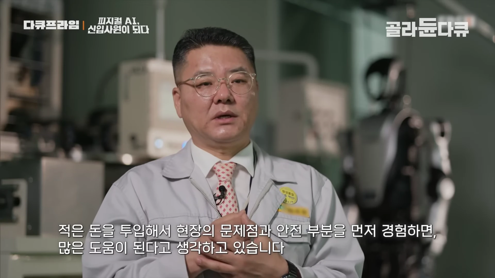 안경을 쓴 남성이 작업복을 입고 인터뷰를 하고 있으며, 뒤쪽으로 휴머노이드 로봇의 실루엣이 보인다.
