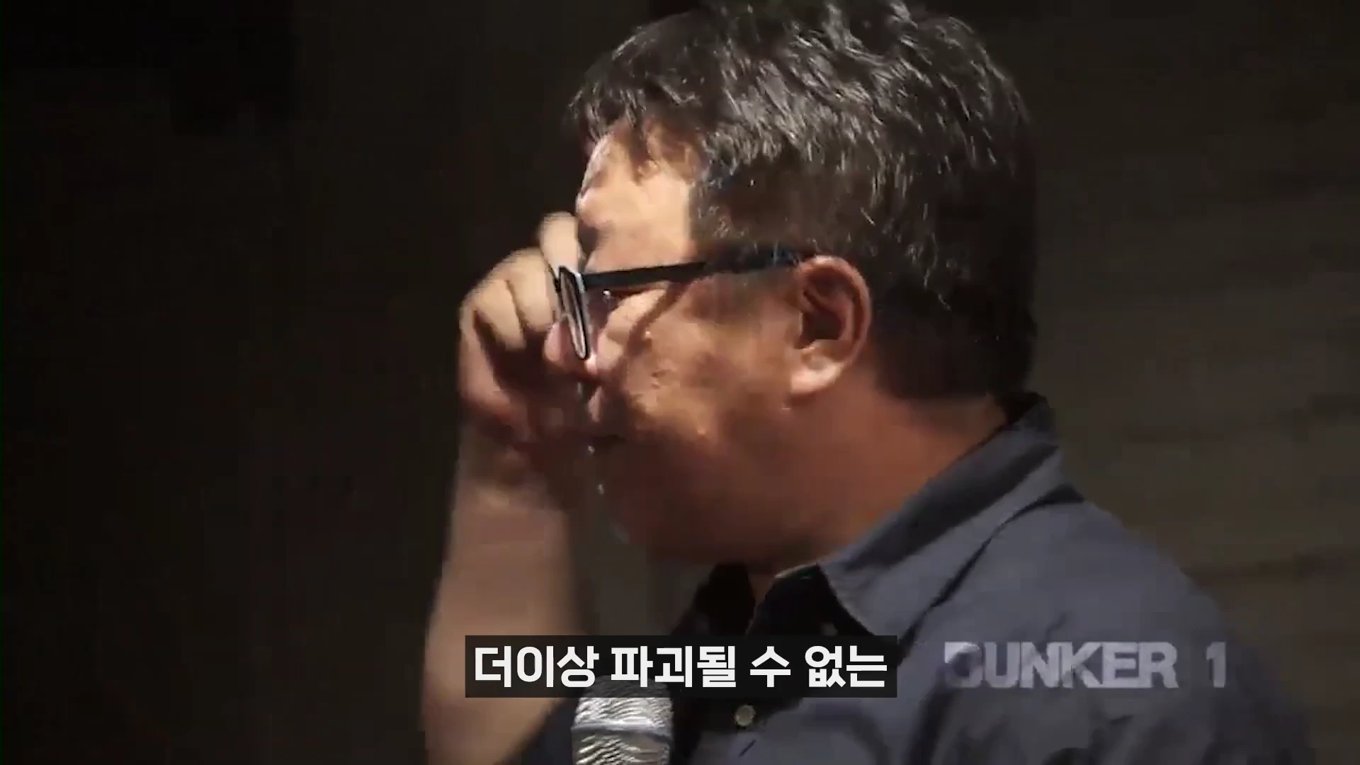 안경을 쓴 중년 남성이 어두운 조명 아래에서 마이크를 잡고 강연하는 모습