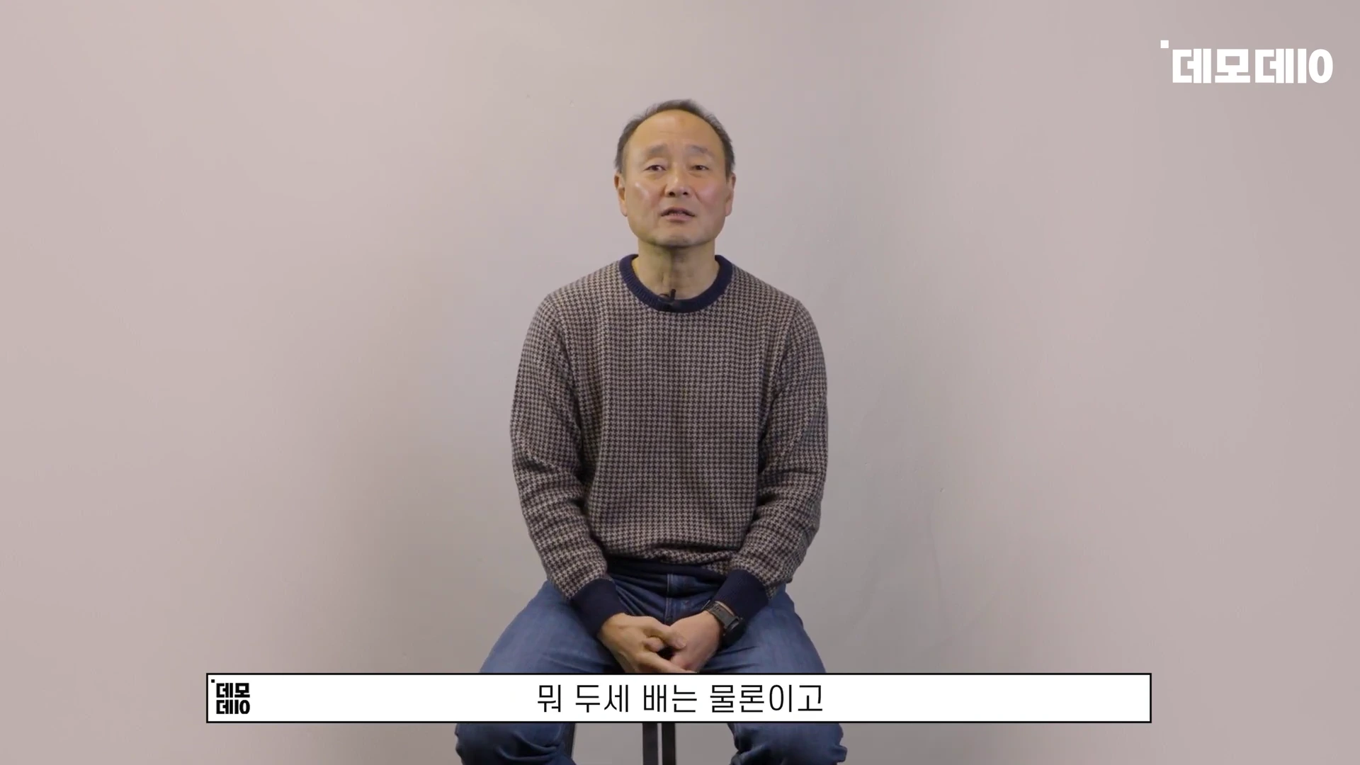 회색 니트를 입은 중년 남성이 의자에 앉아 정면을 바라보며 이야기하고 있는 모습
