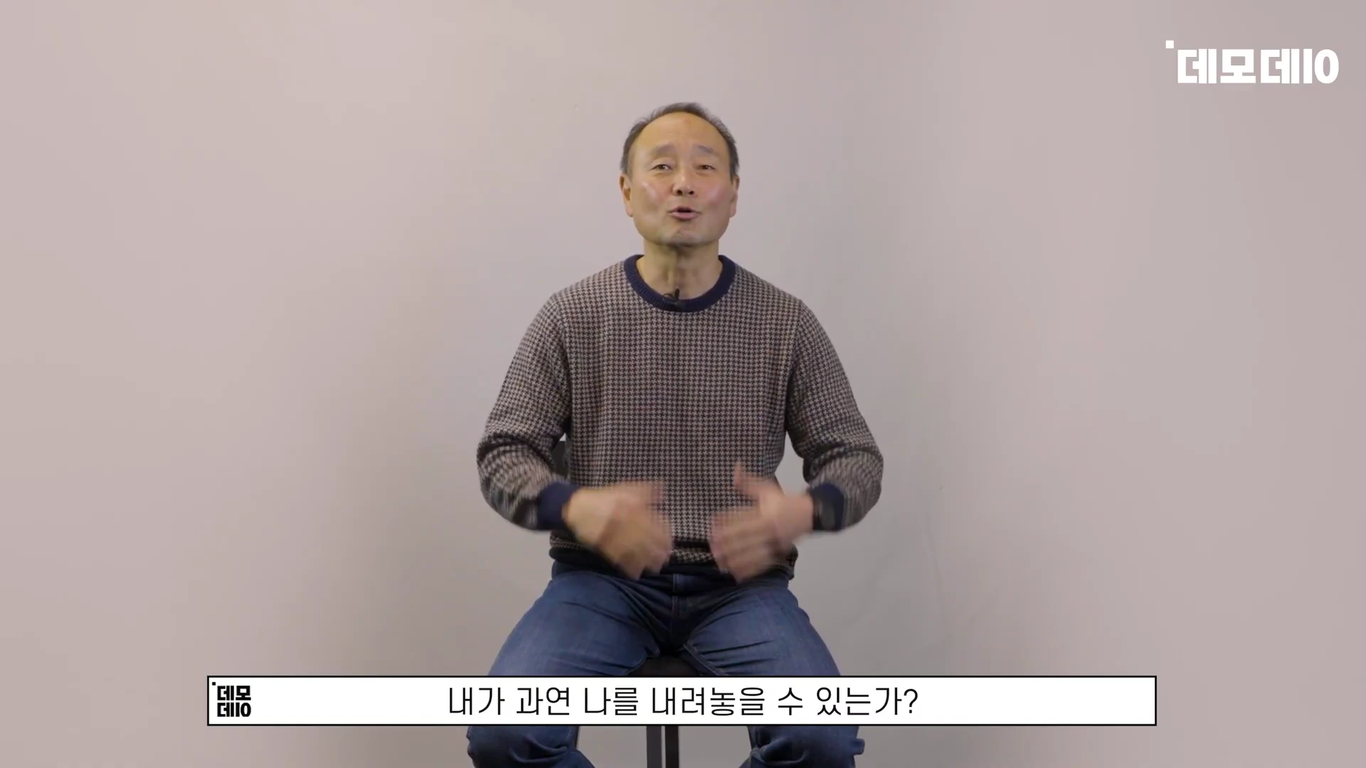 회색 패턴 니트를 입은 중년 남성이 밝은 배경 앞에서 앉아 이야기하고 있는 모습
