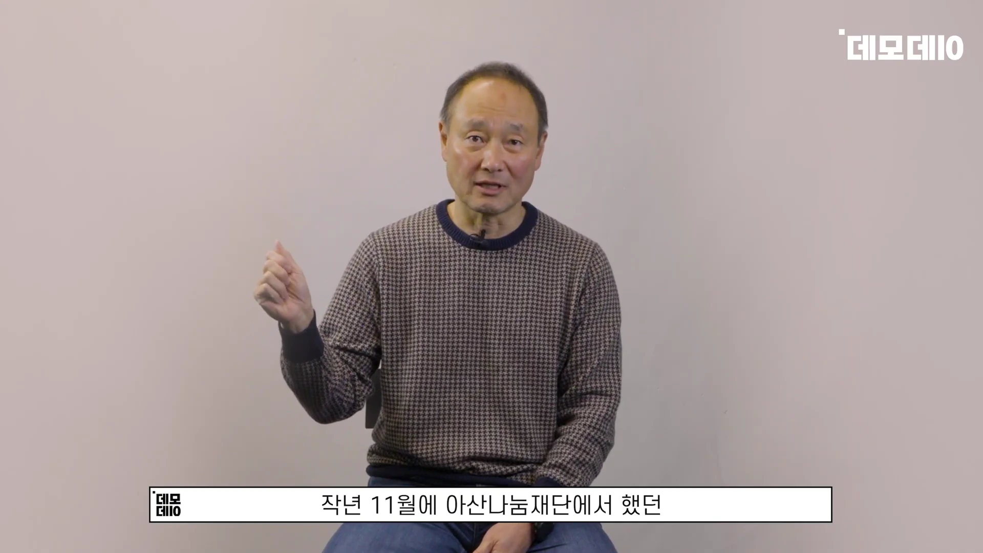 체크무늬 니트를 입은 중년 남성이 밝은 배경 앞에서 손짓하며 이야기하고 있다.