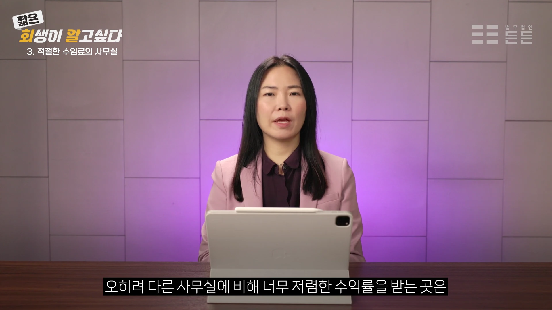 분홍색 재킷을 입은 여성이 책상 앞에 앉아 태블릿을 보며 이야기하는 모습