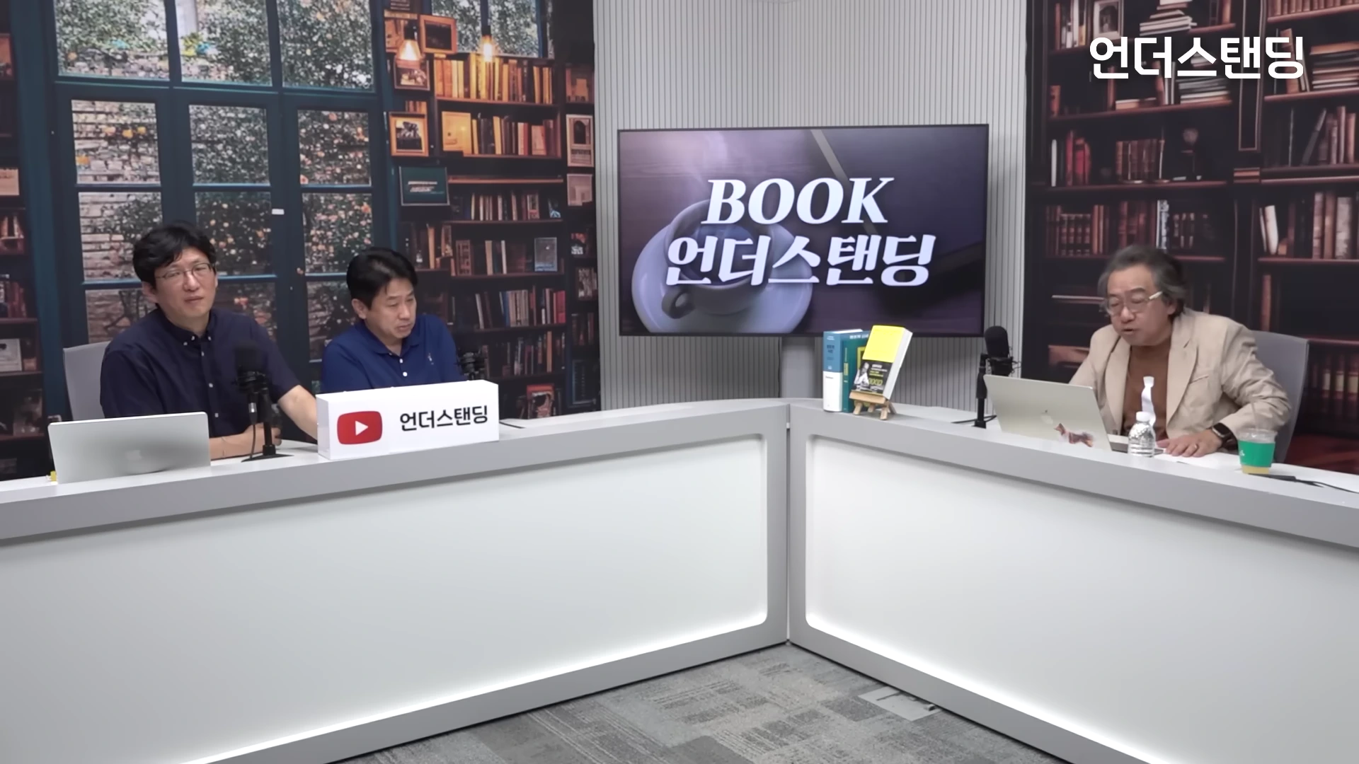 서재 배경의 스튜디오에서 세 명의 남성이 책상에 앉아 대화를 나누는 모습