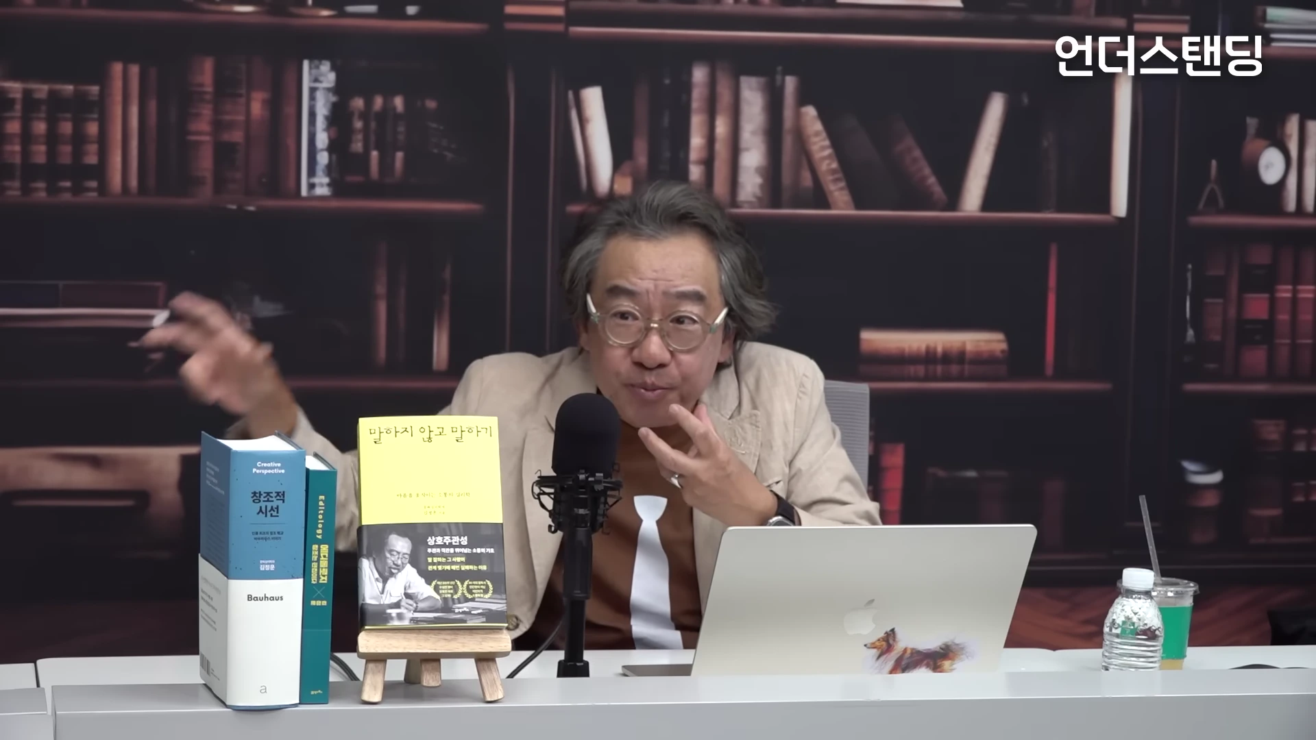 서재 배경의 스튜디오에서 안경을 쓴 중년 남성이 마이크 앞에서 손짓하며 대화하고 있고, 책상 위에는 책들이 놓여 있다.
