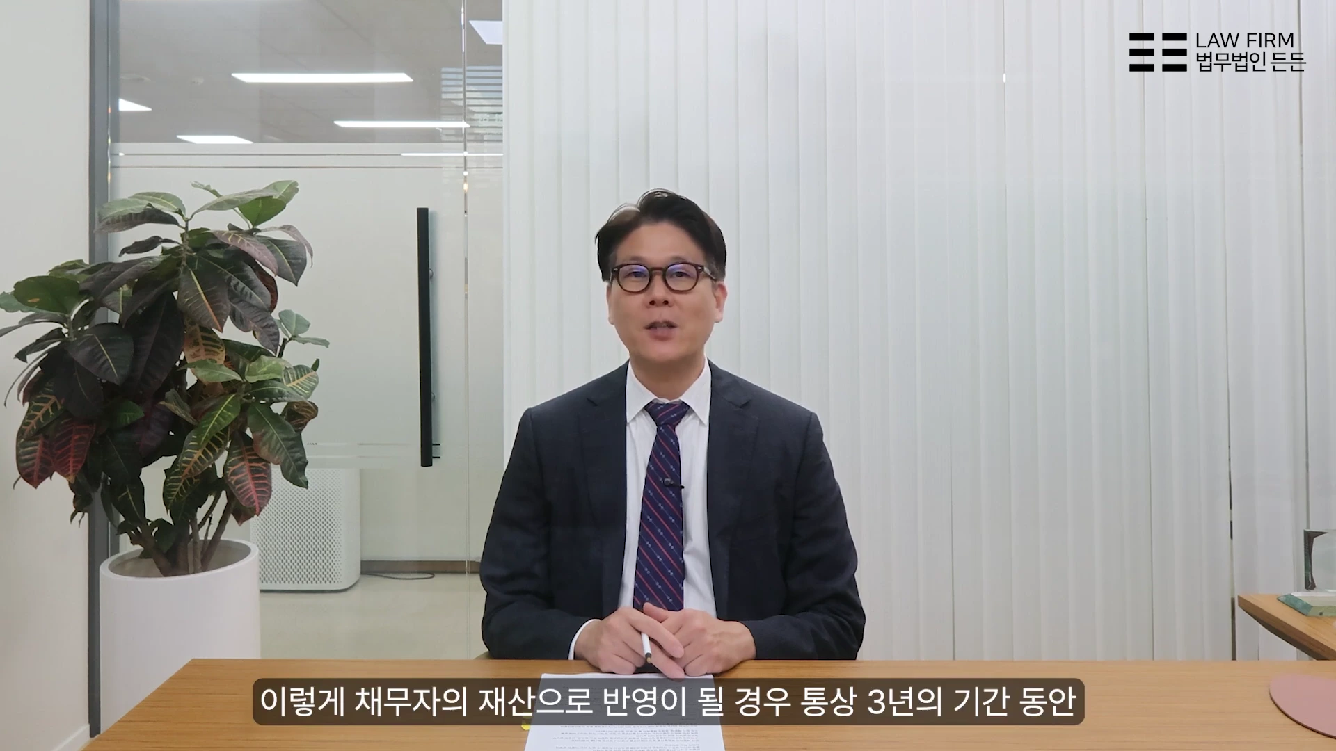 안경을 쓴 남성 변호사가 사무실 책상에 앉아 정면을 보며 이야기하고 있는 모습