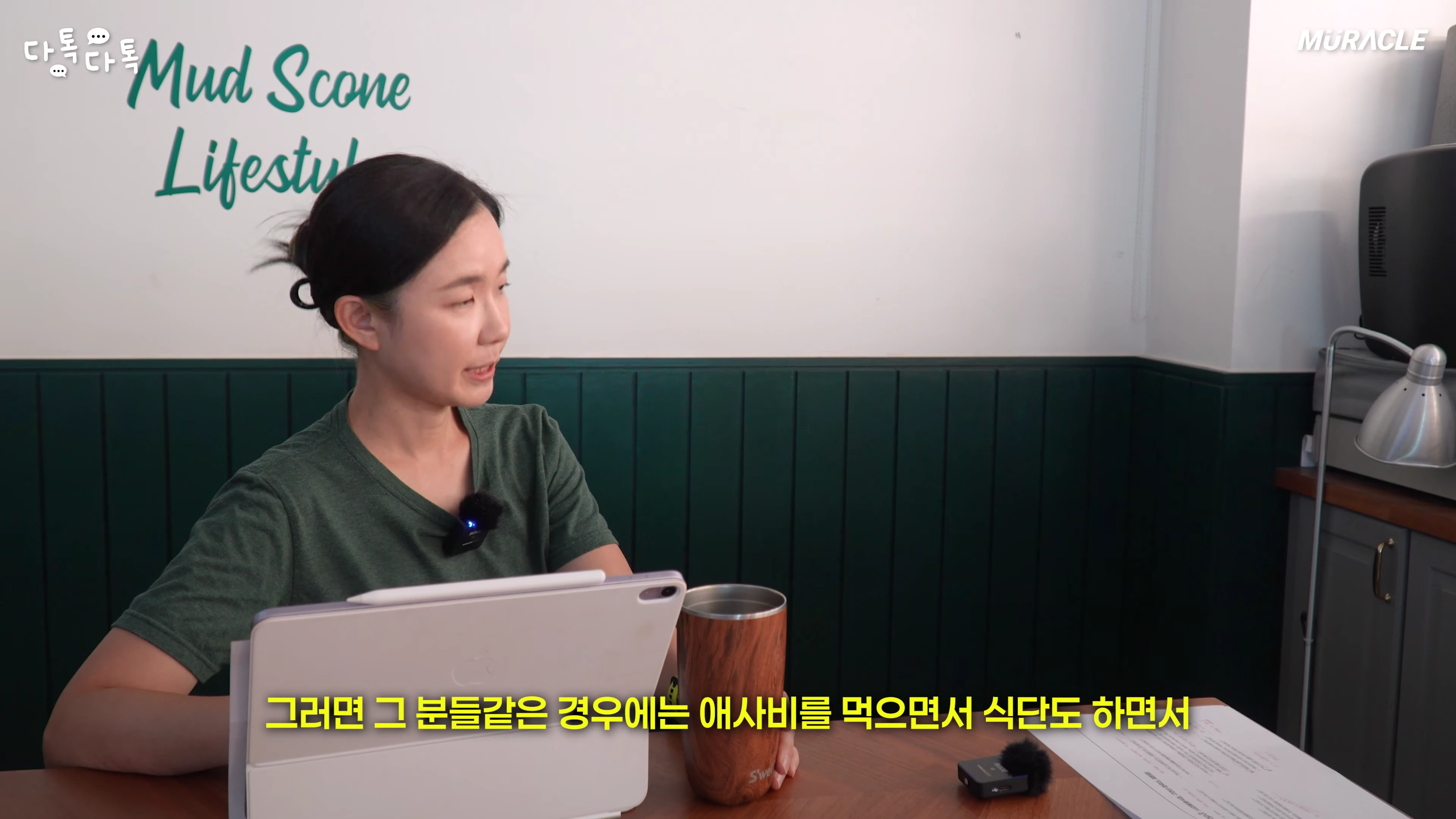 녹색 벽을 배경으로 테이블에 앉아 태블릿을 보며 이야기하는 여성