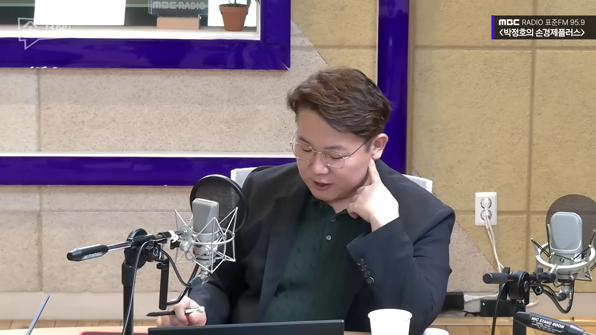 안경을 쓴 남성이 라디오 스튜디오에서 마이크 앞에 앉아 자료를 보며 이야기하고 있다.
