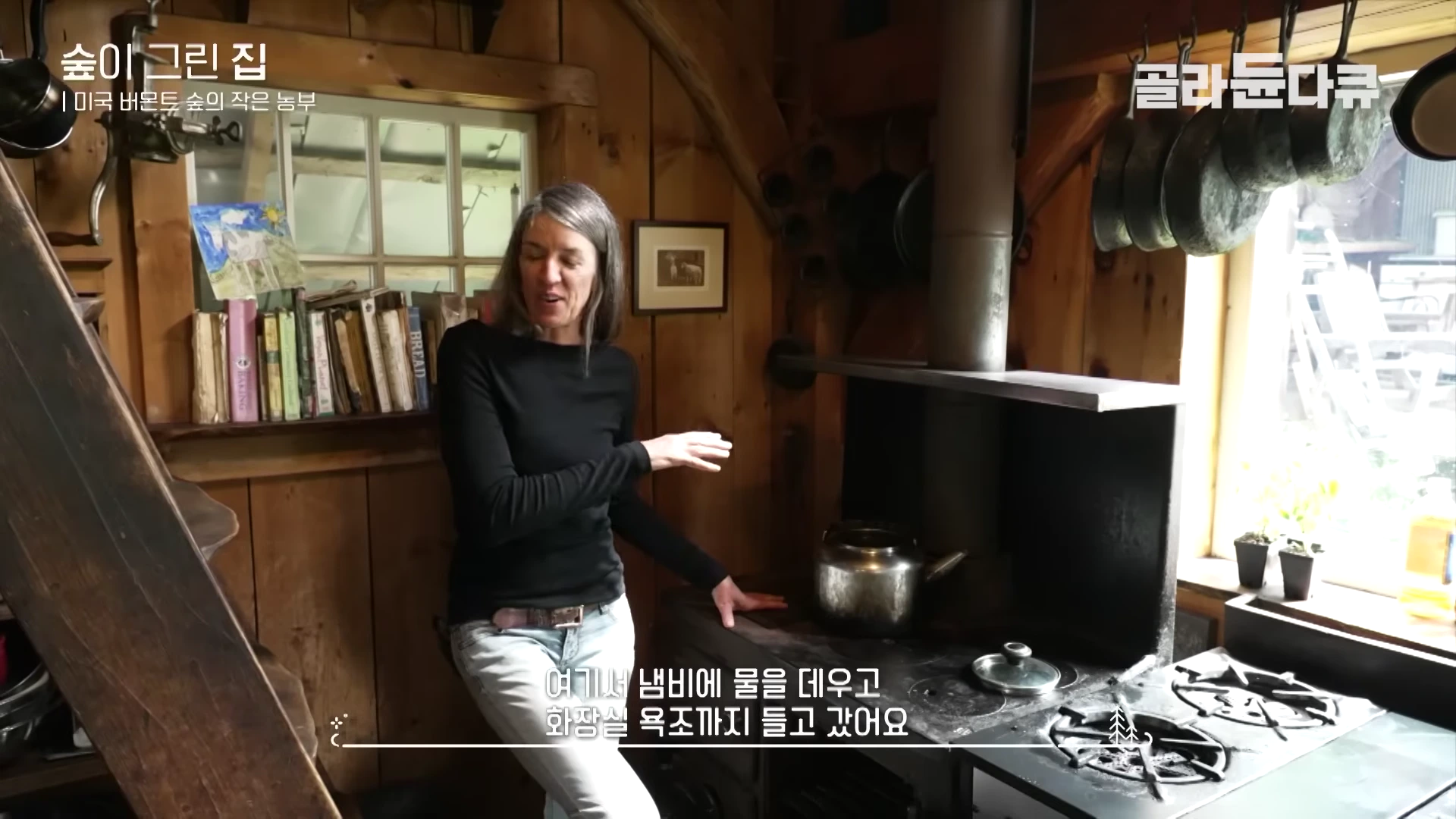 나무 벽으로 된 주방에서 한 여성이 옛날식 화목 난로 옆에 서서 손짓하며 이야기하고 있다.