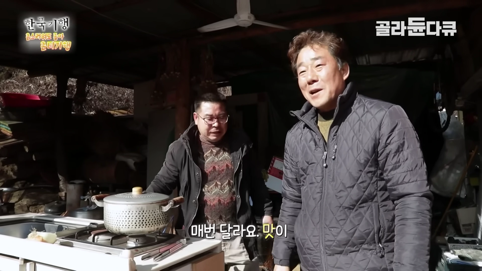 야외 주방에서 두 남자가 김치찌개가 담긴 냄비를 앞에 두고 대화를 나누고 있다.