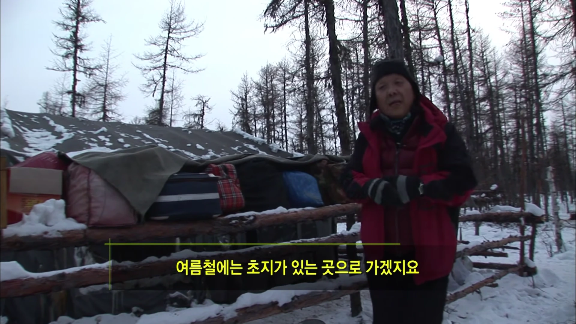 눈 덮인 숲속에서 붉은 조끼를 입은 남성이 인터뷰하고 있으며, 뒤편에는 짐이 쌓인 천막 구조물이 보인다.