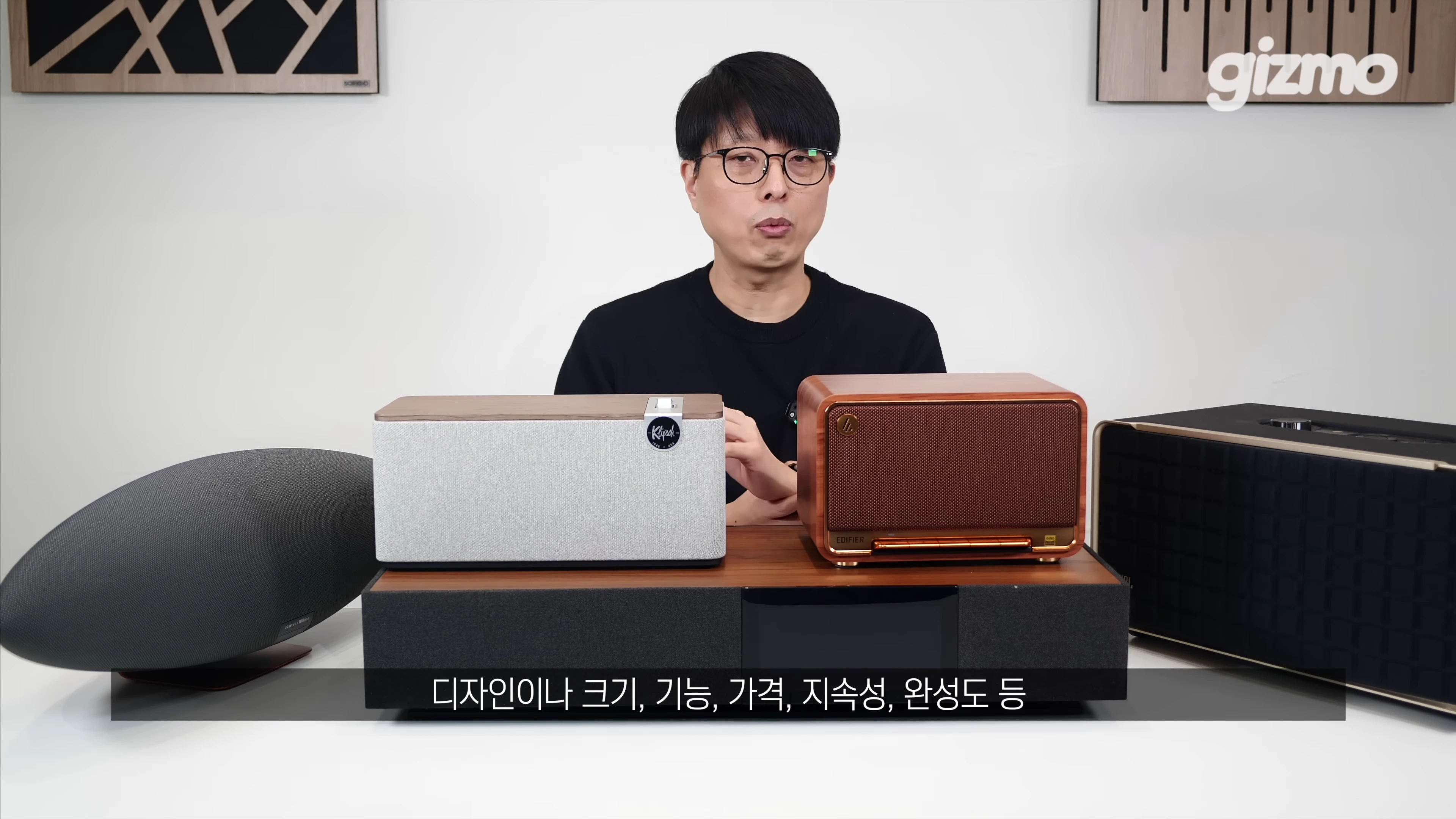 안경을 쓴 남성이 다양한 디자인의 올인원 홈 스피커들이 놓인 테이블 앞에 앉아 설명하고 있다.