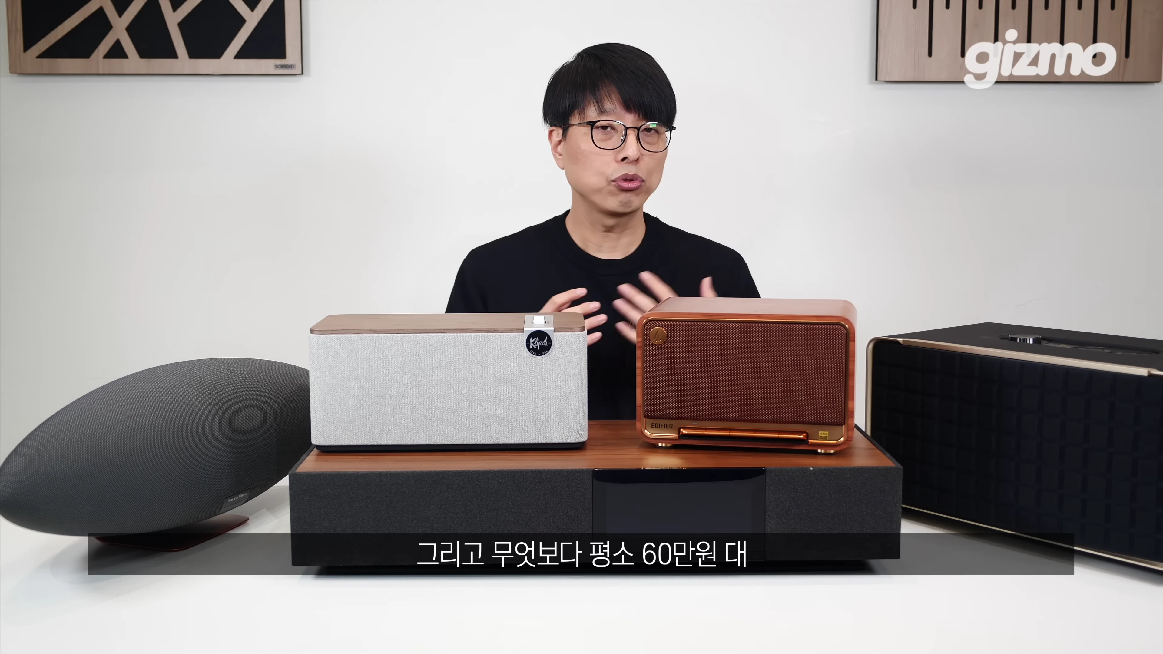 안경을 쓴 남성이 다양한 디자인의 스피커들을 앞에 두고 설명하는 스튜디오 영상 화면
