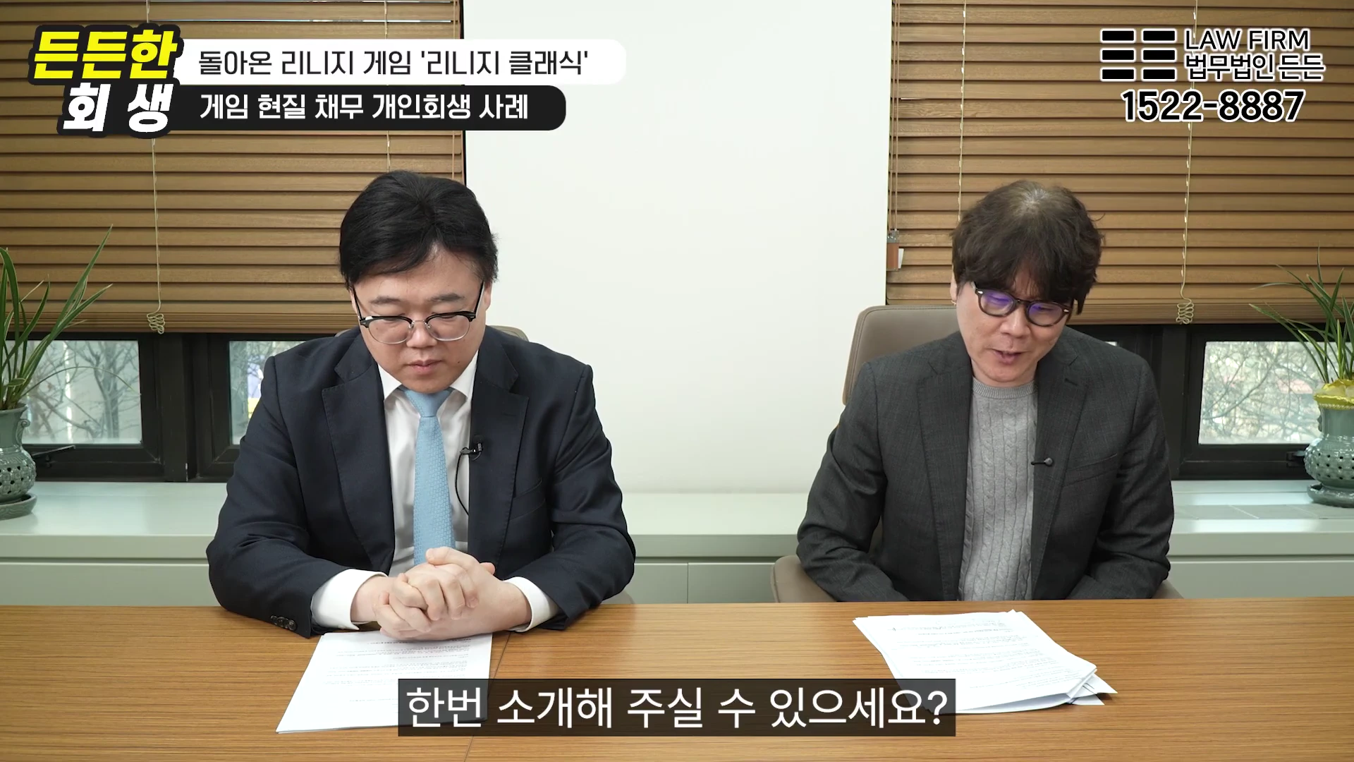정장을 입은 두 명의 남성이 사무실 책상에 앉아 서류를 보며 대화하는 모습