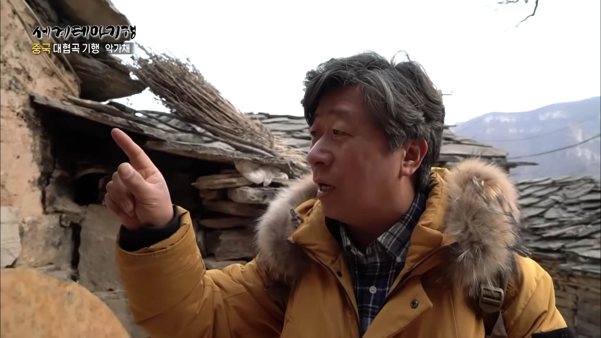 노란색 패딩을 입은 중년 남성이 돌로 쌓은 집 앞에서 손가락으로 어딘가를 가리키며 이야기하고 있다.