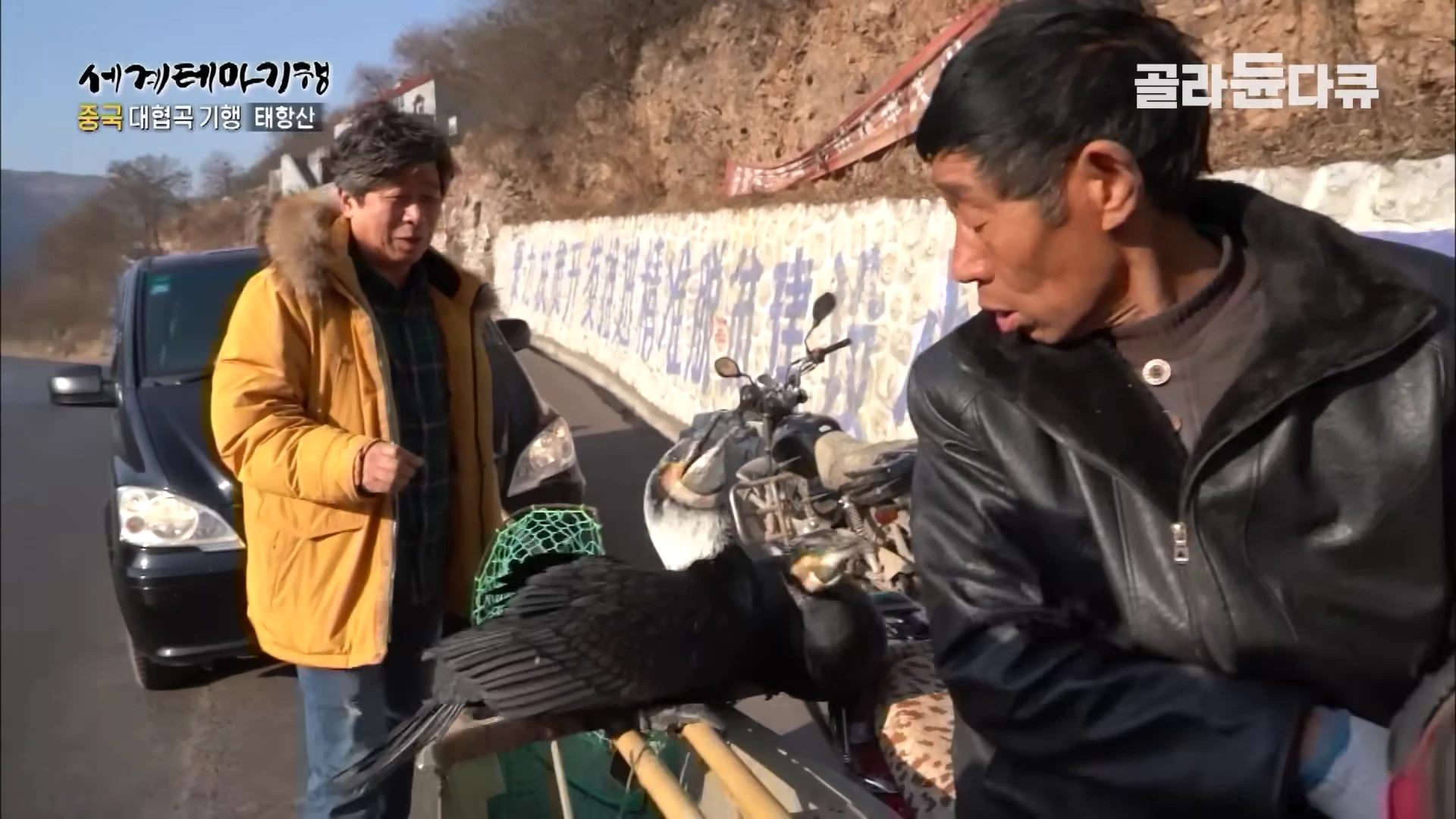 가마우지를 데리고 있는 어부와 노란 점퍼를 입은 남성이 길가에서 대화하는 모습