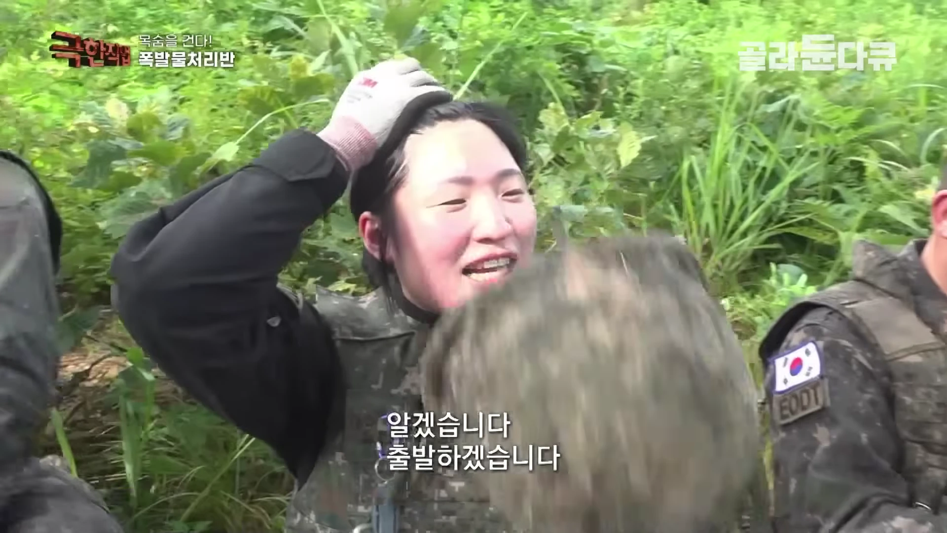 군복을 입은 대원이 숲속에서 헬멧을 고쳐 쓰며 땀을 닦고 있는 모습