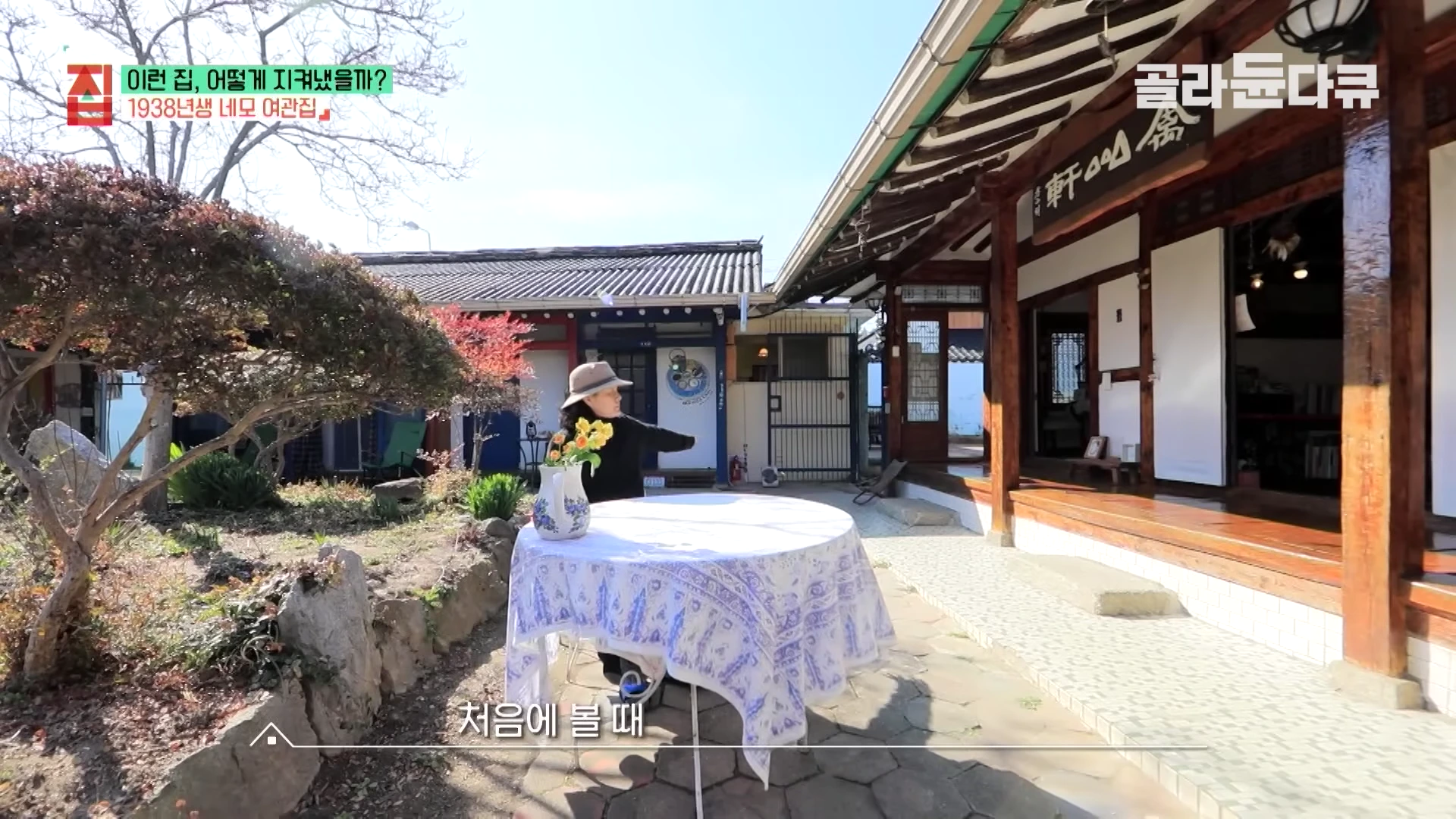 한옥 마당에 모자를 쓴 여성이 서 있고, 앞에는 꽃병이 놓인 둥근 테이블이 있다.
