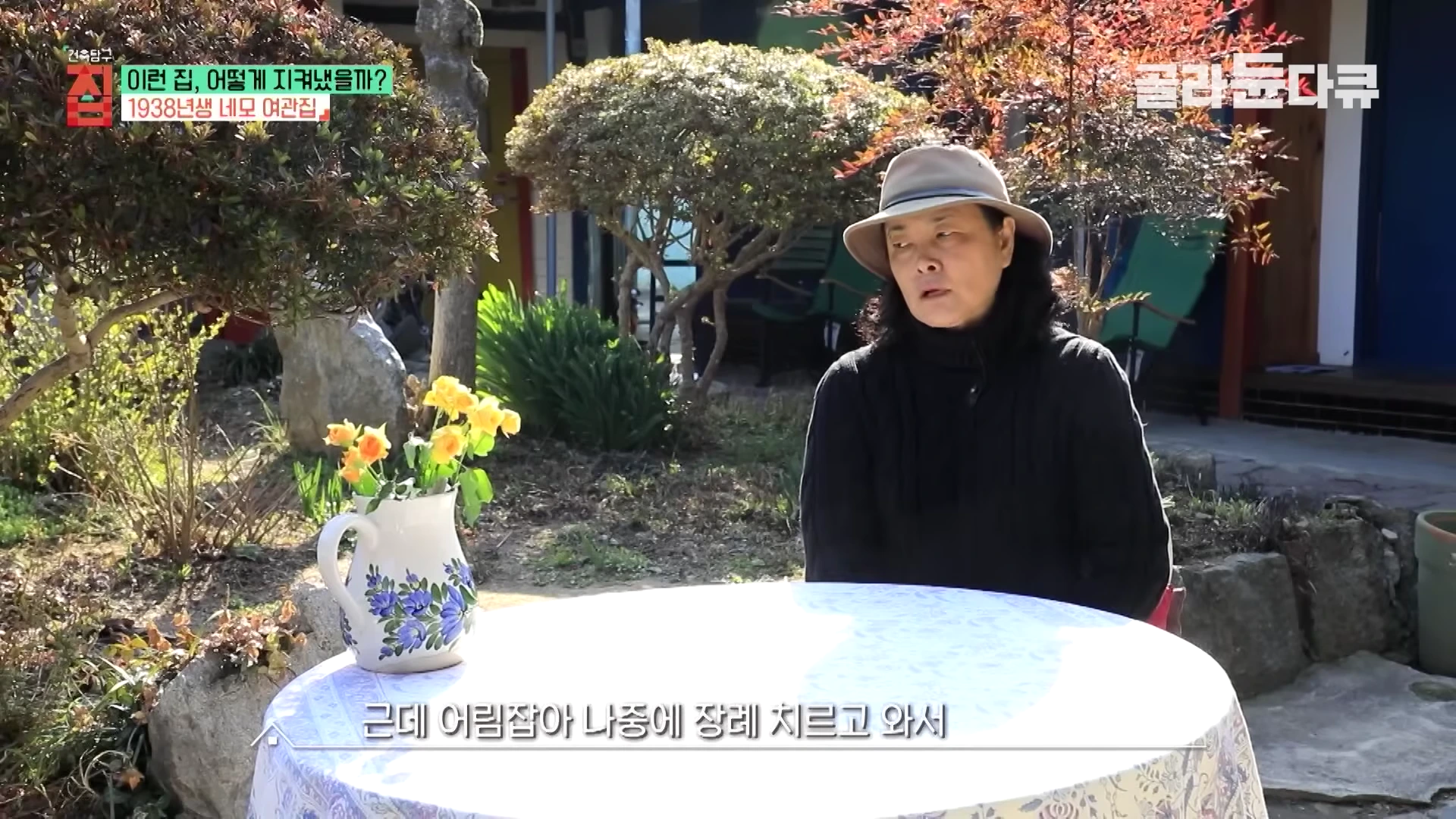 햇살이 비치는 정원에서 모자를 쓴 여성이 테이블 앞에 앉아 인터뷰하는 모습
