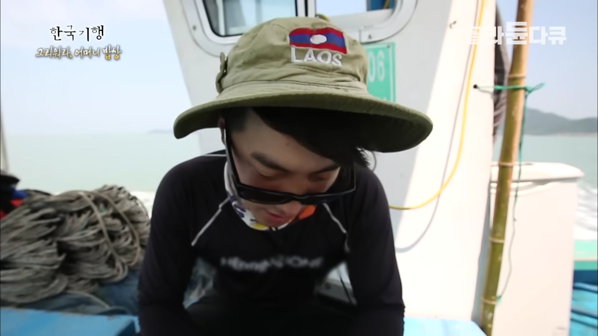 라오스 로고가 새겨진 모자와 선글라스를 쓴 청년이 배 위에서 아래를 내려다보고 있다.