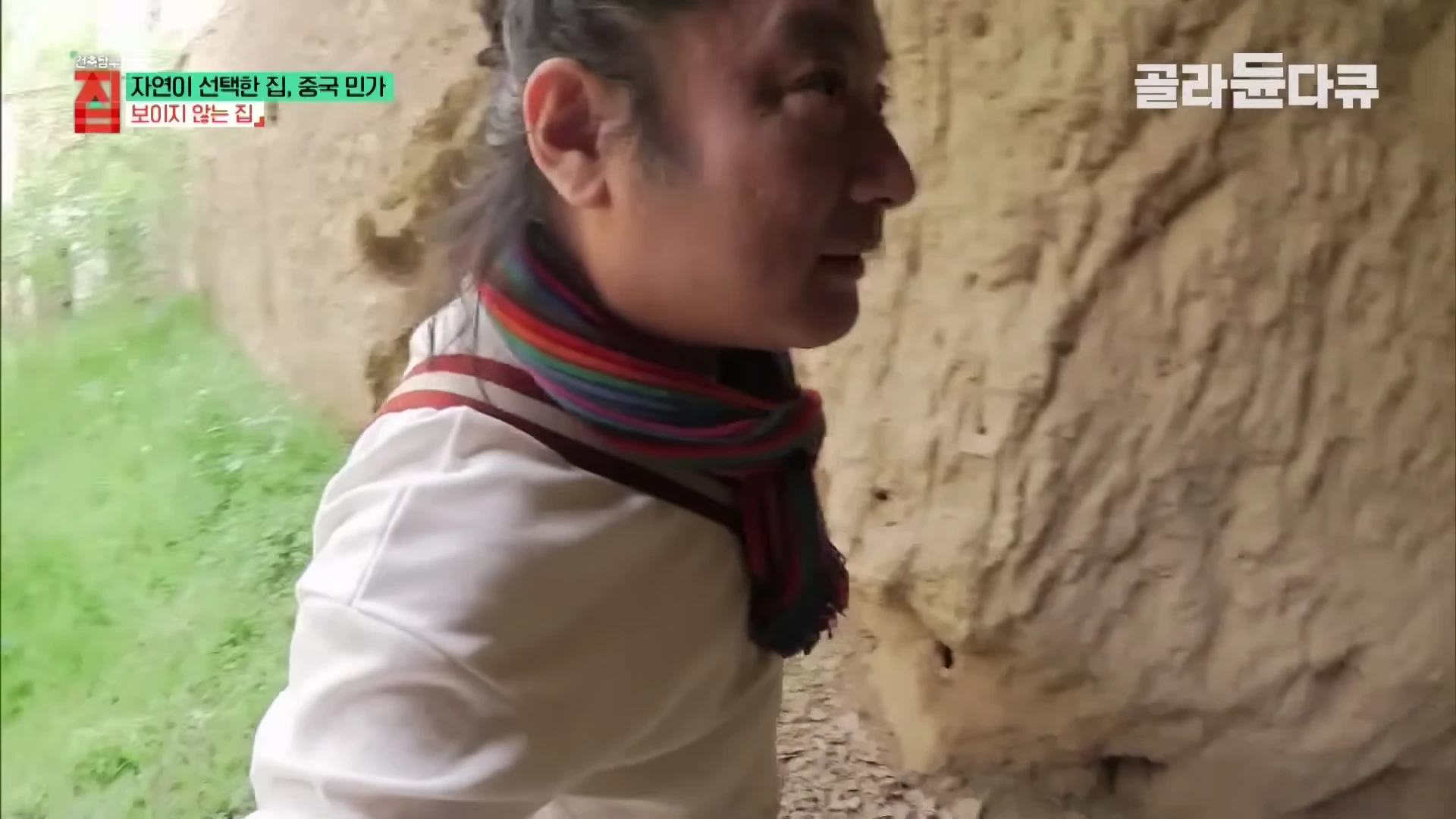 스카프를 두른 남성이 황토 벽으로 된 동굴 형태의 집 내부를 살펴보고 있다.