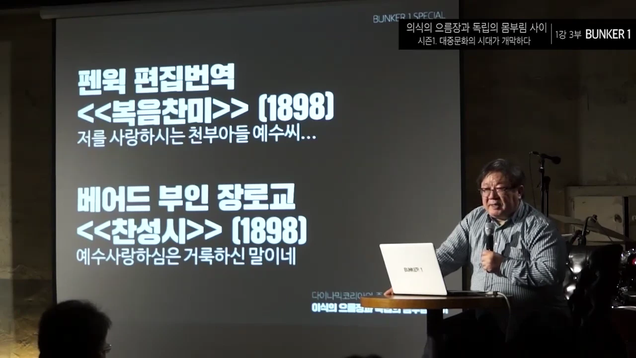 강연자가 스크린 앞에 앉아 마이크를 잡고 있으며, 스크린에는 1898년 발행된 찬송가 가사 비교 내용이 적혀 있습니다.