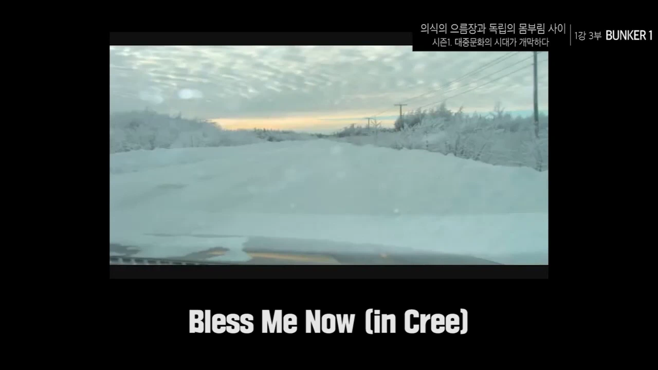 눈 덮인 도로를 달리는 자동차 안에서 촬영한 풍경 영상과 하단에 'Bless Me Now (in Cree)'라는 자막이 표시된 화면.