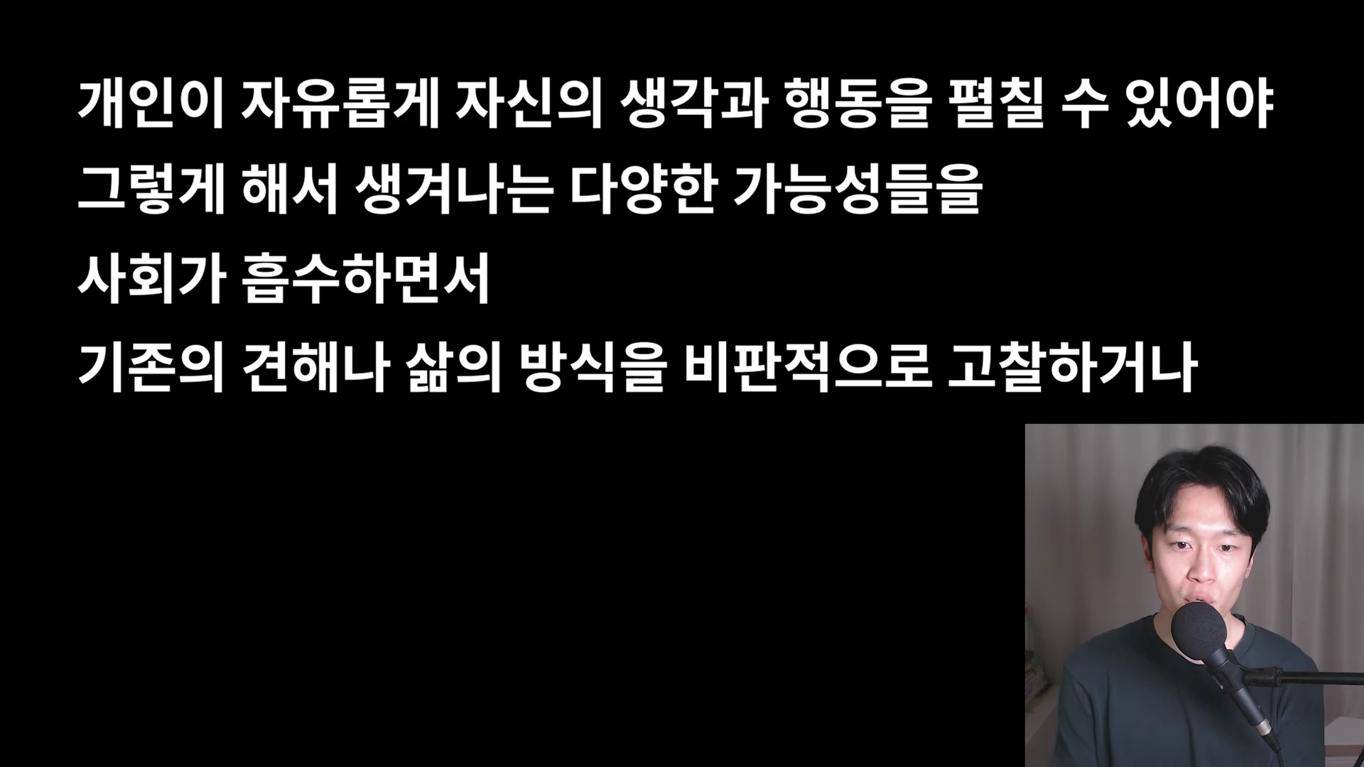 검은 배경에 흰색 자막이 적혀 있고, 오른쪽 하단에 마이크 앞에 앉은 남성이 보이는 영상 화면