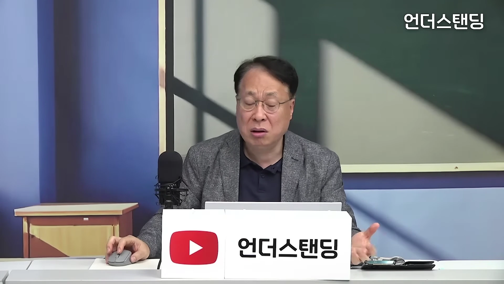 안경을 쓴 중년 남성이 스튜디오 책상에 앉아 마이크 앞에서 손짓하며 이야기를 나누고 있다.