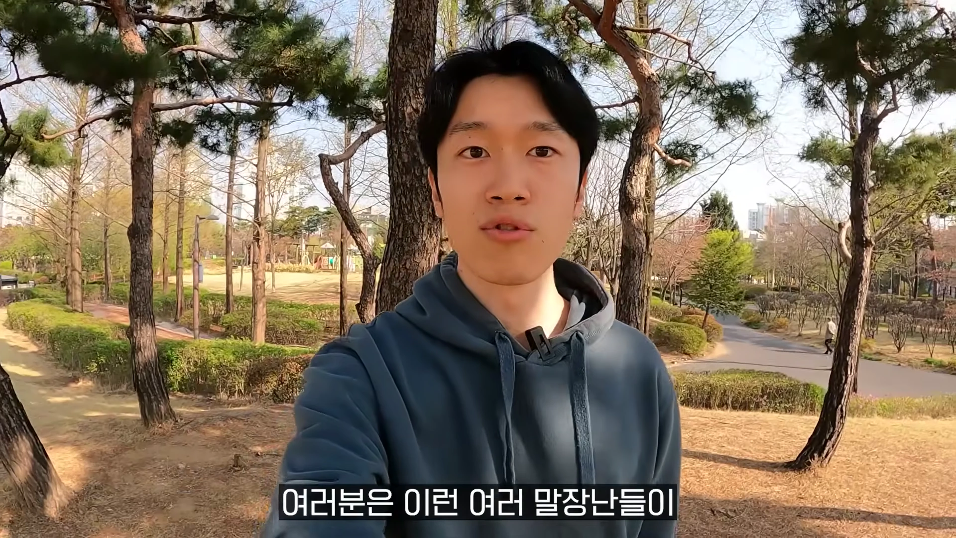 공원 산책로를 배경으로 후드티를 입은 남성이 카메라를 응시하며 이야기하는 모습