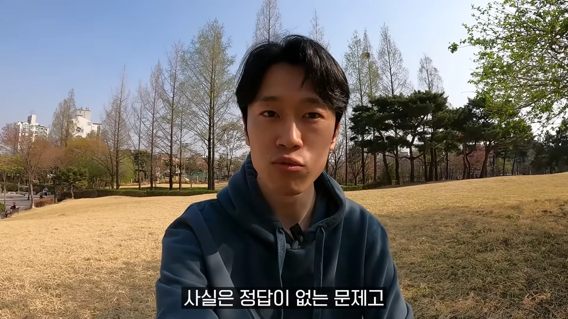 야외 공원을 배경으로 후드티를 입은 남성이 카메라를 응시하며 이야기하는 모습