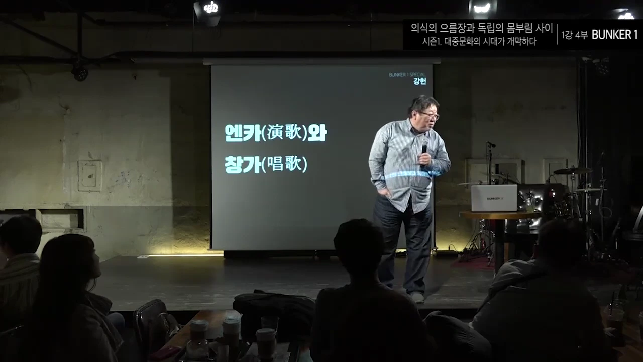 강연자가 어두운 무대 위 스크린 앞에 서서 마이크를 들고 청중을 향해 이야기하고 있다.