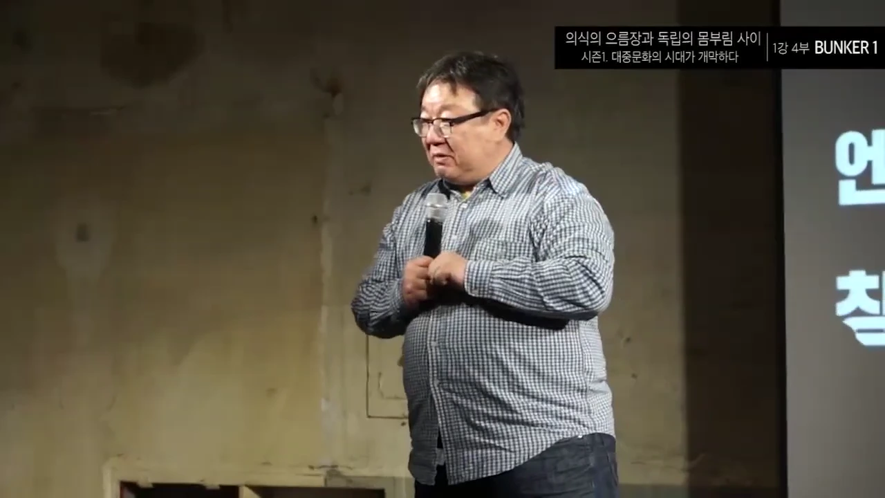 안경을 쓴 중년 남성이 체크무늬 셔츠를 입고 마이크를 든 채 강연하고 있는 모습