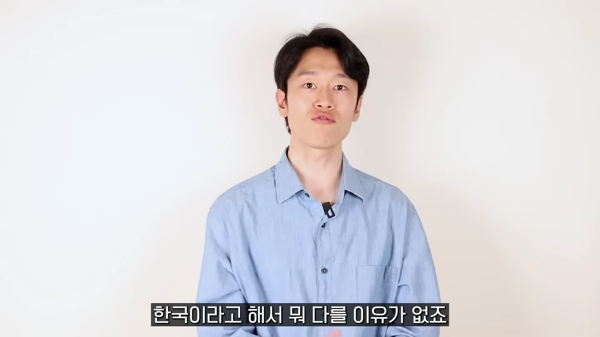흰색 배경 앞에서 하늘색 셔츠를 입고 정면을 응시하며 말하는 남성