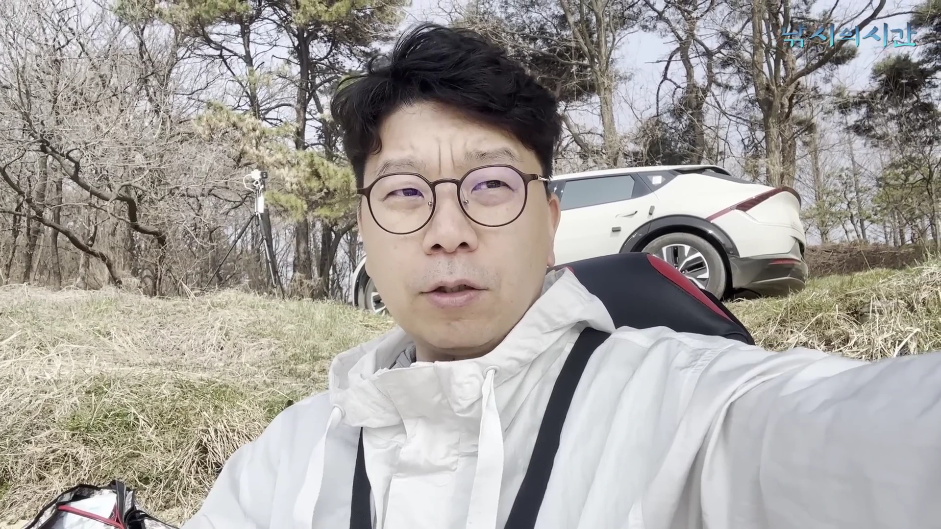 야외 낚시터에 앉아 카메라를 응시하며 이야기하는 안경 쓴 남성