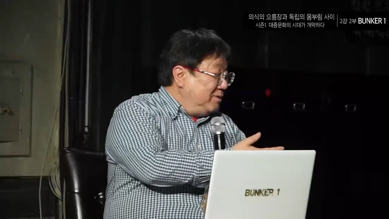 체크무늬 셔츠를 입고 안경을 쓴 중년 남성이 노트북 앞에 앉아 강연하는 모습