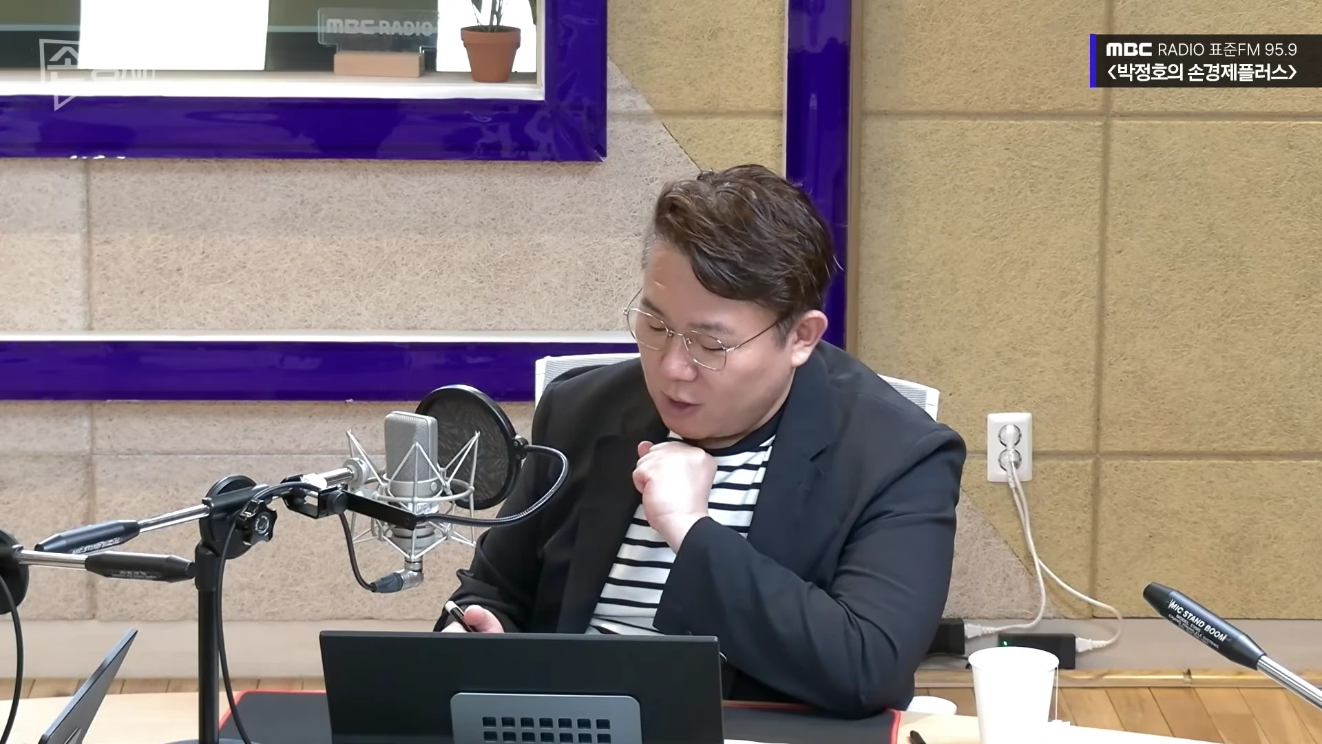 라디오 스튜디오에서 안경을 쓴 남성이 마이크 앞에 앉아 노트북을 보며 이야기하고 있다.