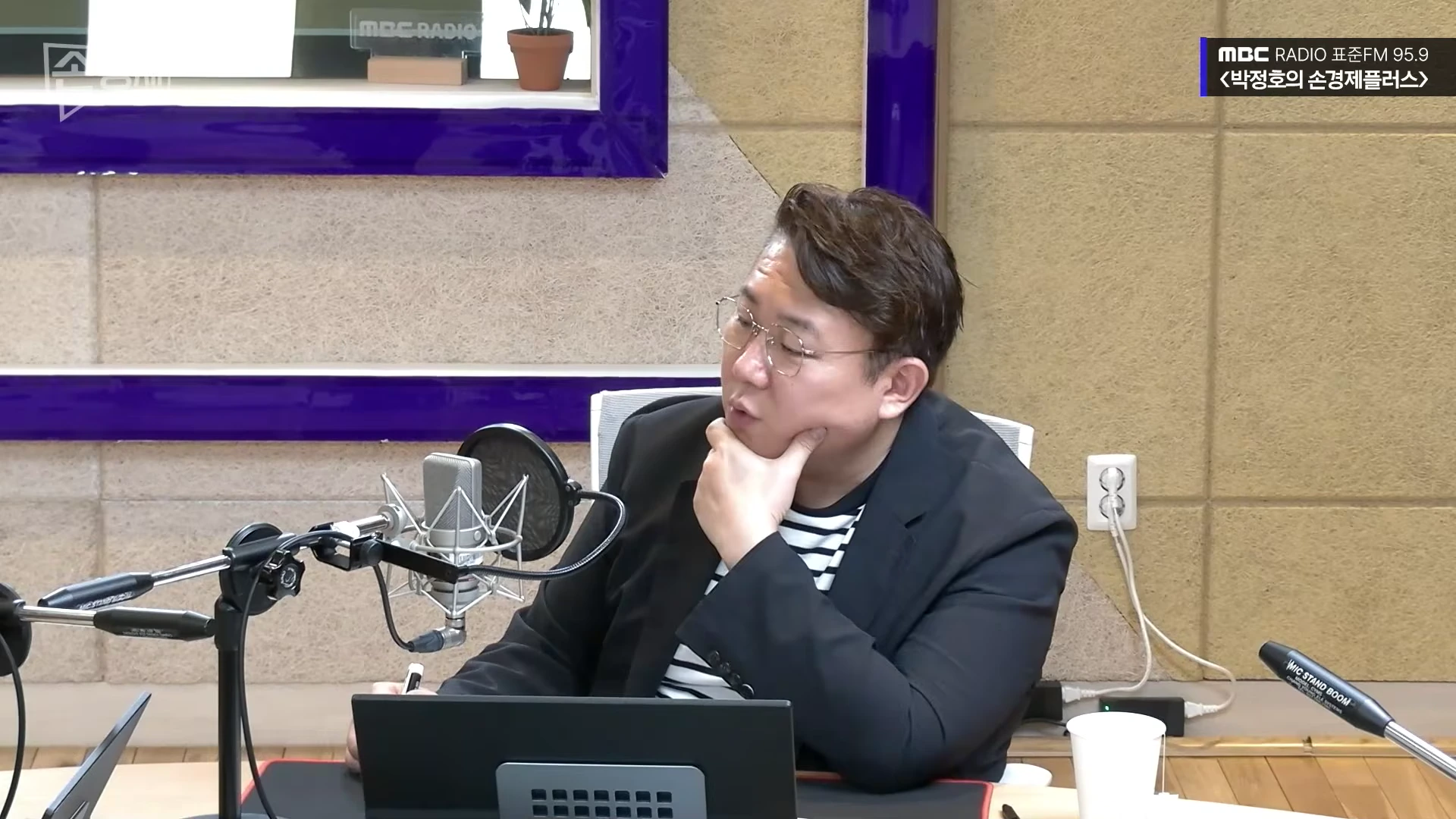안경을 쓴 남성이 라디오 스튜디오에서 마이크 앞에 앉아 턱을 괴고 생각에 잠긴 모습