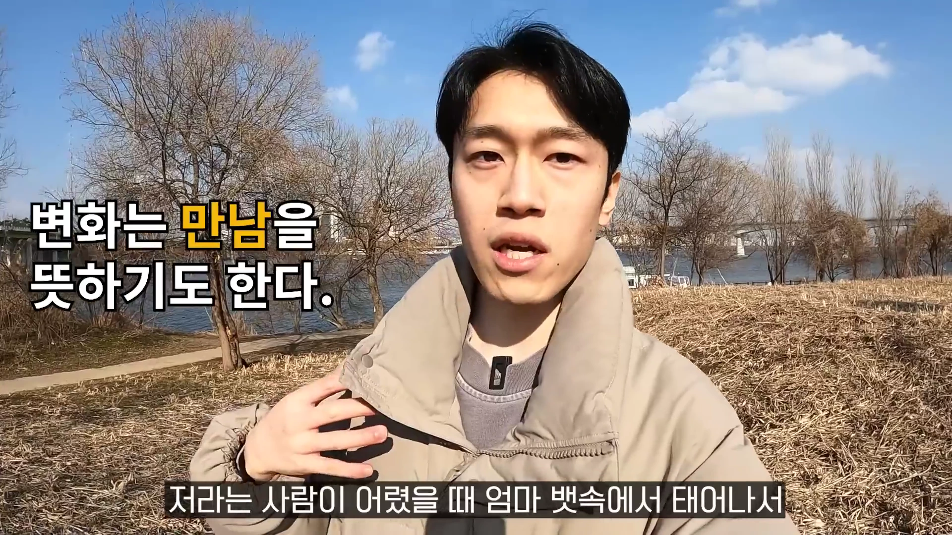 야외 강변을 배경으로 베이지색 패딩을 입은 남성이 카메라를 보며 이야기하고 있다.