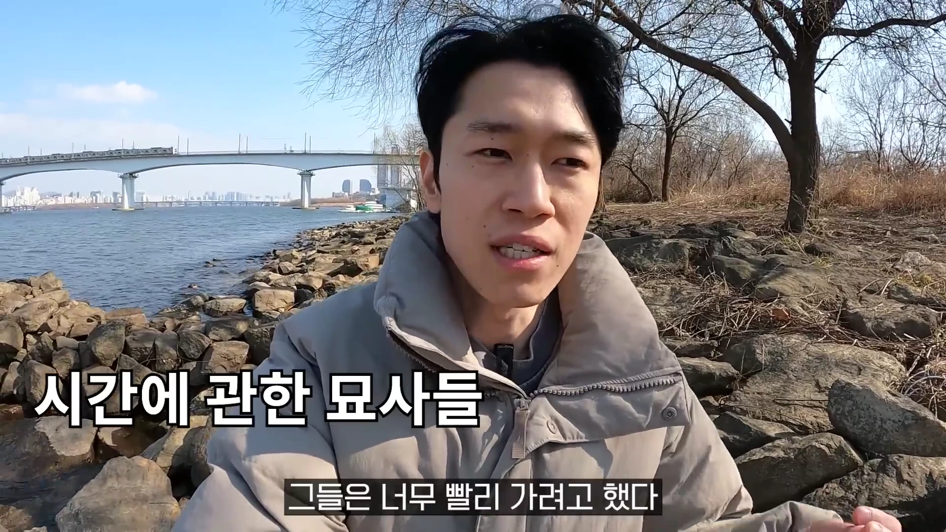 강변 돌밭에 앉아 이야기하는 남성과 화면 하단에 표시된 자막