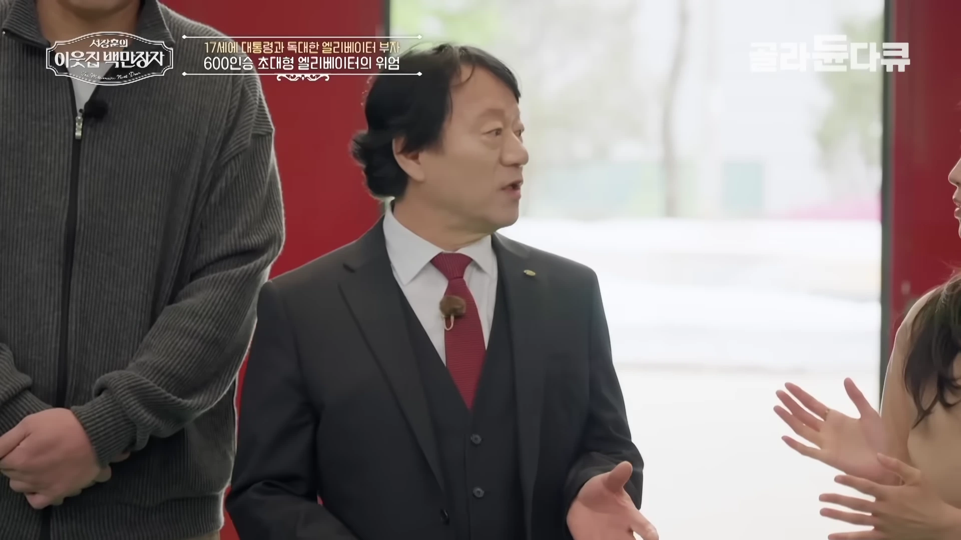 정장을 입은 중년 남성이 인터뷰하며 손짓으로 설명하고 있고, 양옆으로 다른 출연자들의 모습이 일부 보인다.
