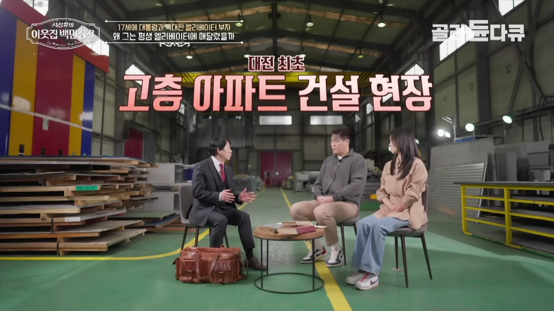 넓은 공장 내부에서 세 사람이 의자에 앉아 대화를 나누고 있으며, 화면 상단에는 '대전 최초 고층 아파트 건설 현장'이라는 자막이 적혀 있습니다.