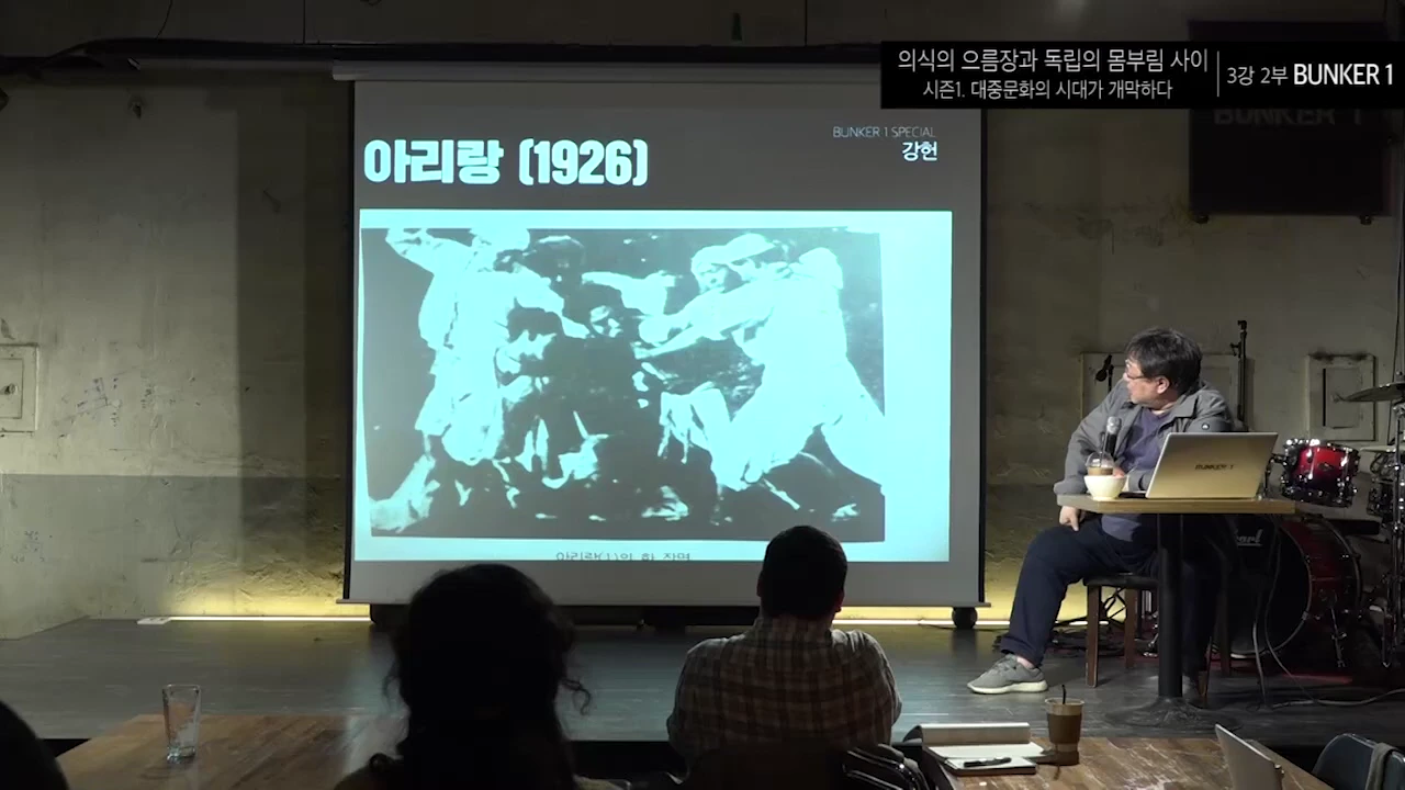 강연자가 스크린에 투사된 1926년 영화 아리랑의 흑백 스틸컷을 가리키며 청중에게 설명하고 있다.