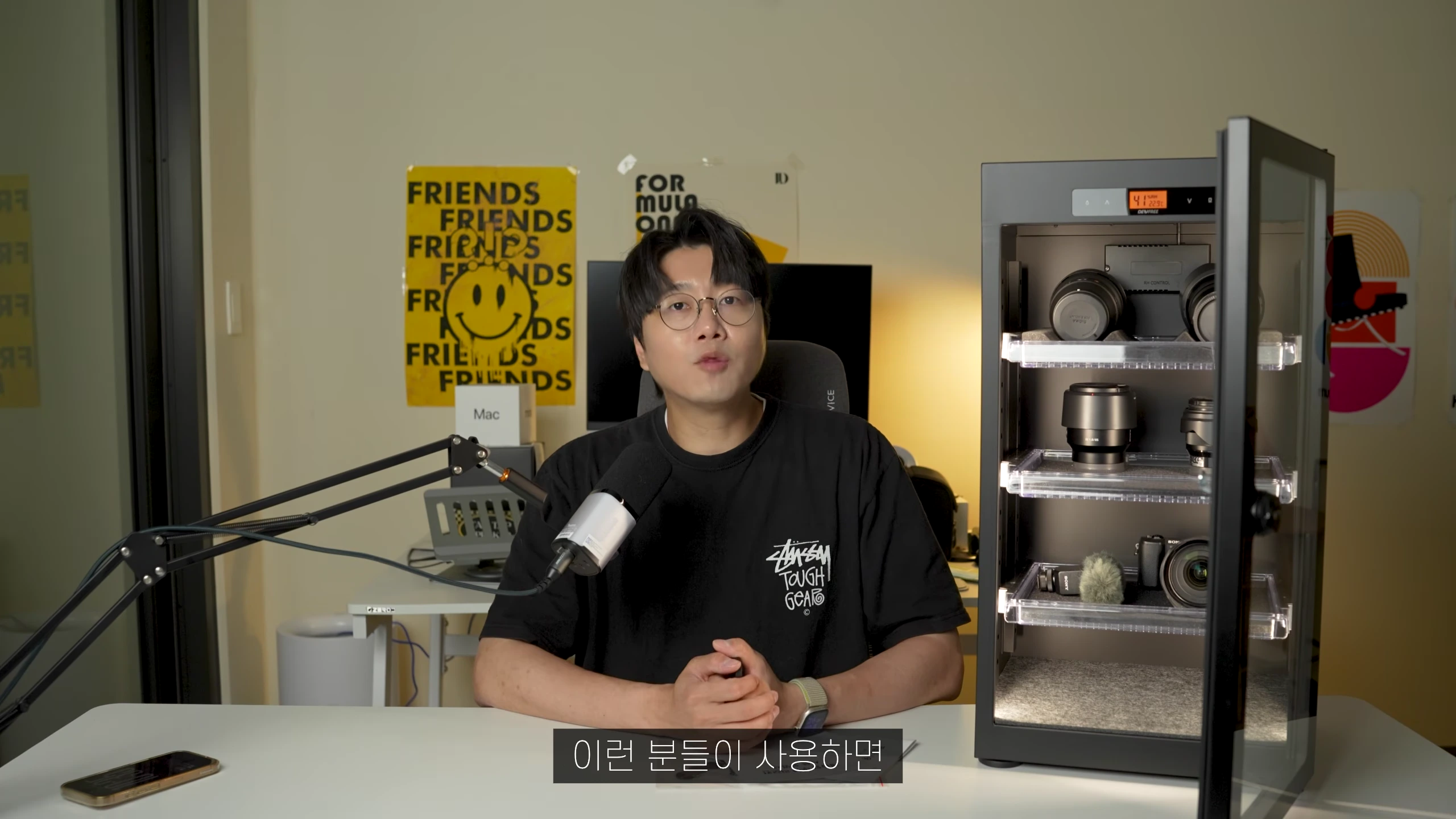 안경을 쓴 남성이 마이크 앞에 앉아 있고 옆에는 카메라 장비가 보관된 제습함이 놓여 있다.